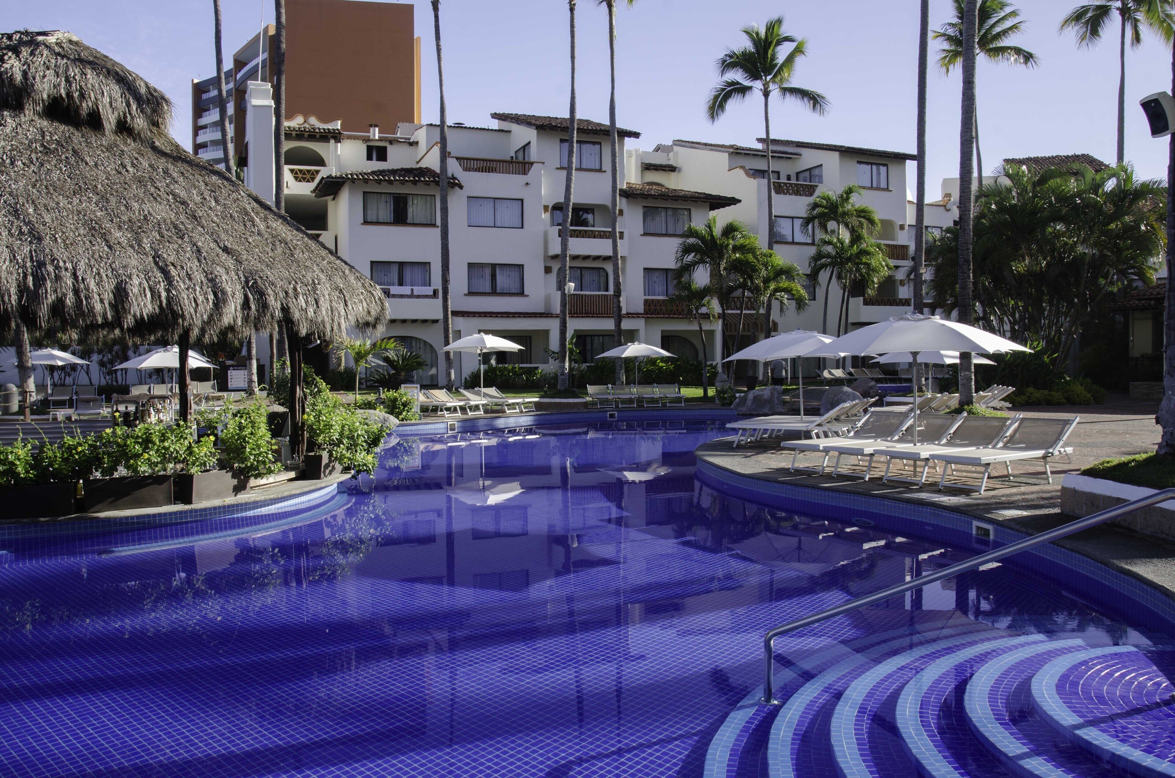 Una piscina con sillones de descanso y sombrillas, rodeada de palmeras y un edificio con balcones.