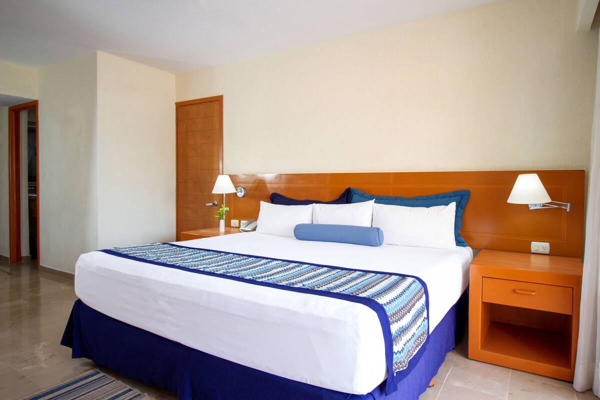 Una habitación de hotel con una cama grande, cabecera de madera, dos mesitas de noche con lámparas, un tocador y una puerta que da a otra habitación.