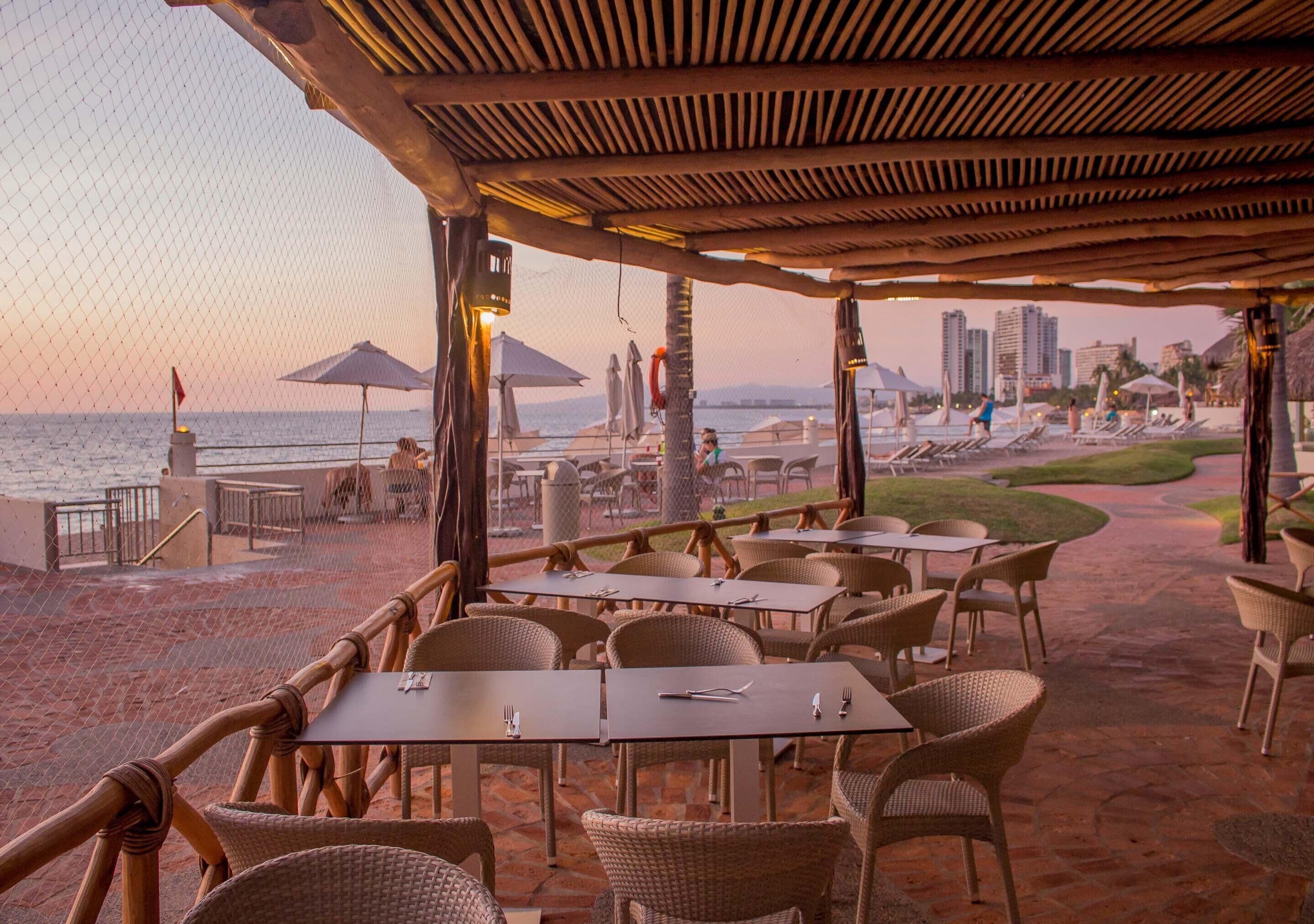 Un área de comedor al aire libre con mesas y sillas, con vistas a una playa y al horizonte de la ciudad.