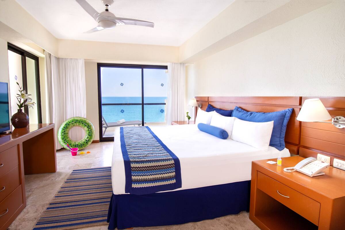 Habitación de hotel con una cama grande, una mesita de noche de madera, un escritorio, una silla, un ventanal con vista al océano y un ventilador de techo.