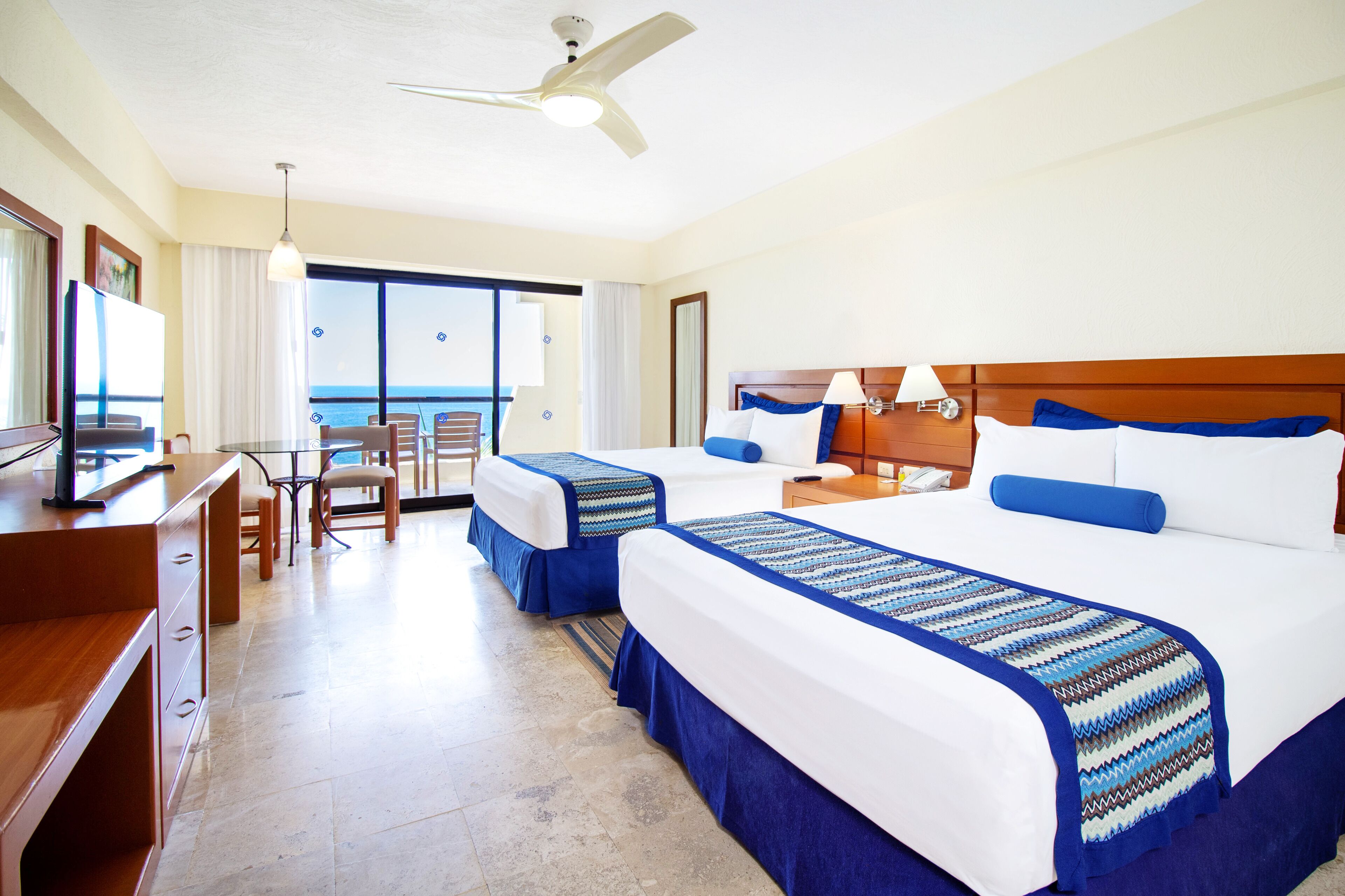 A hotel room with two beds, a desk, and a chair. There is a balcony with a view of the ocean.