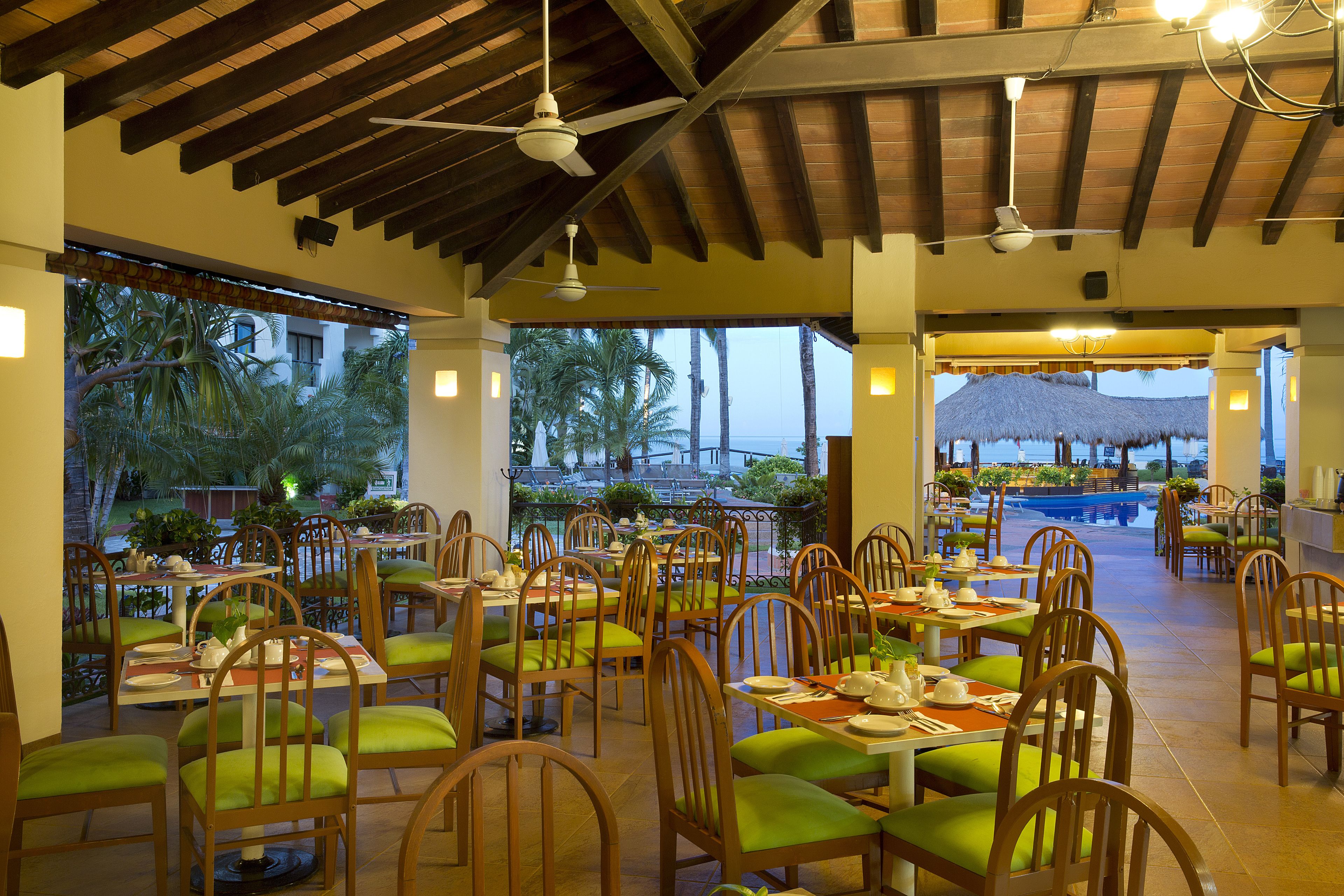 A restaurant with wooden chairs and tables, large windows, and a view of palm trees and a pool.
