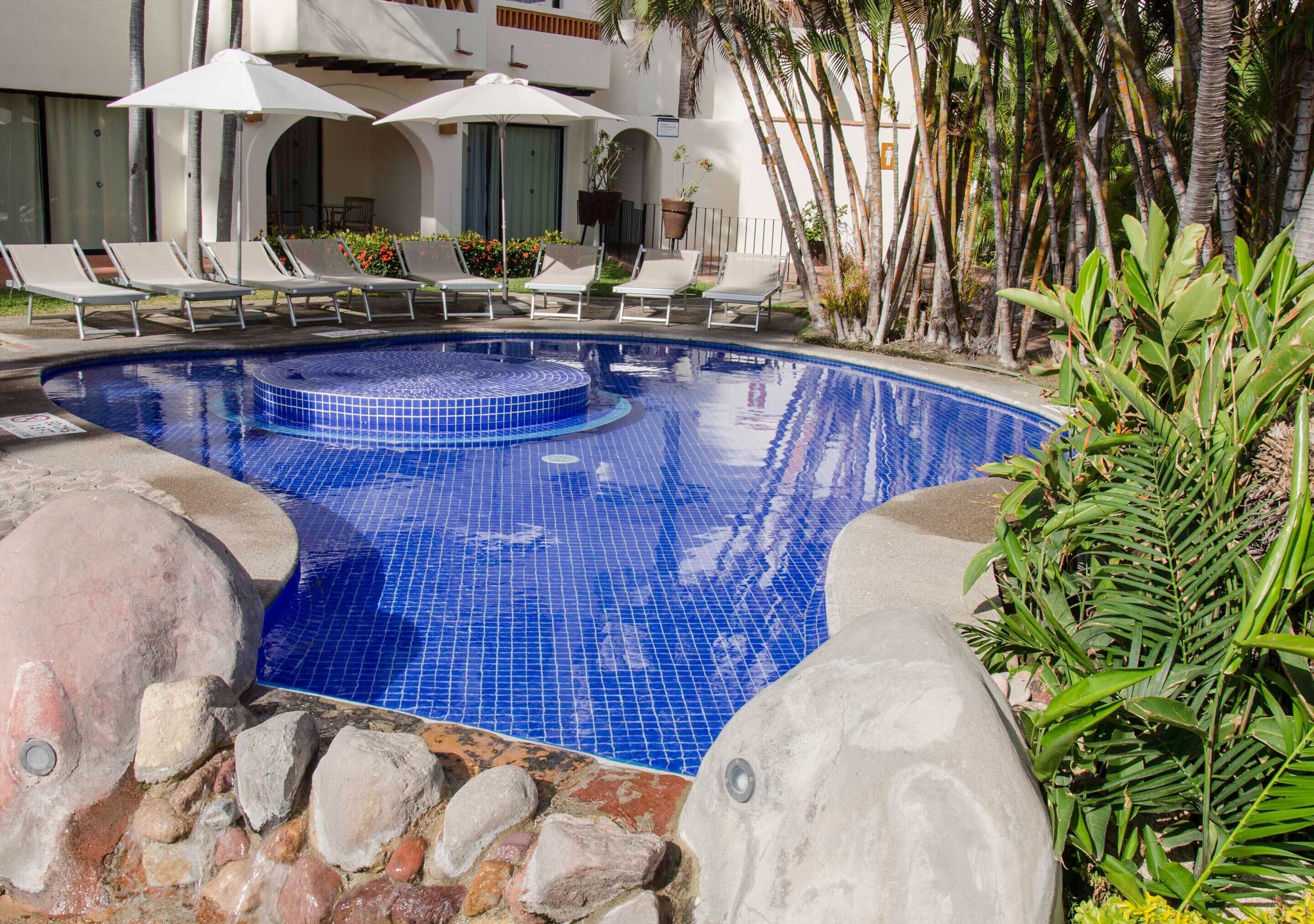 A swimming pool with lounge chairs and umbrellas, surrounded by tropical plants and a building with balconies.