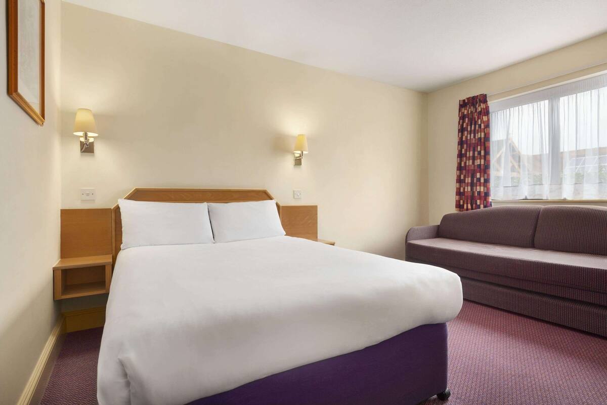 A hotel room with a bed, a sofa, two wall-mounted lamps, and a window with curtains.
