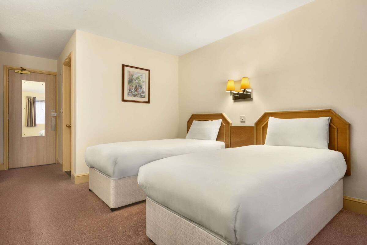 A hotel room with two beds, a door, a framed picture on the wall, and a wall-mounted lamp.