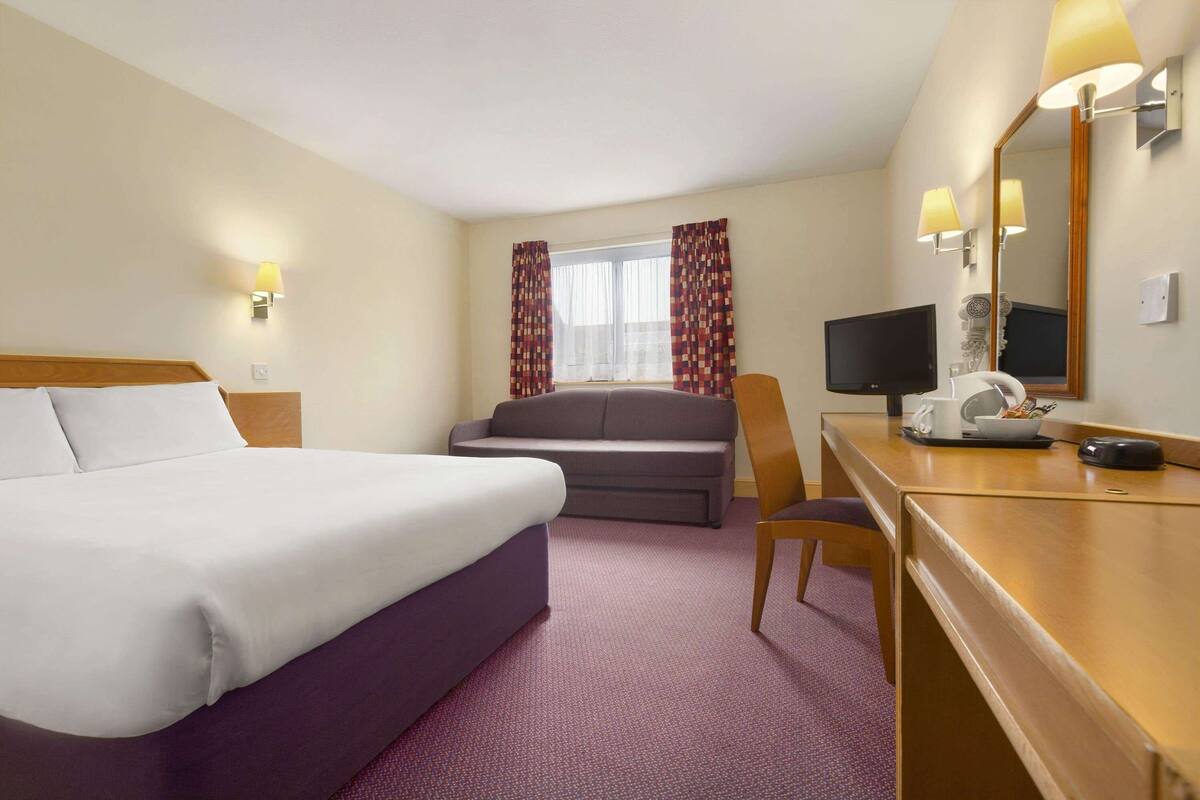A hotel room with a bed, a sofa, a desk with a chair, a television, and a mirror.