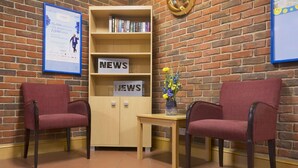 A reception area with two red chairs, a wooden bookshelf with 'NEWS' signs, a small table with a vase, and a brick wall. - Days Inn by Wyndham Sevenoaks Clacket Lane (Westerham)