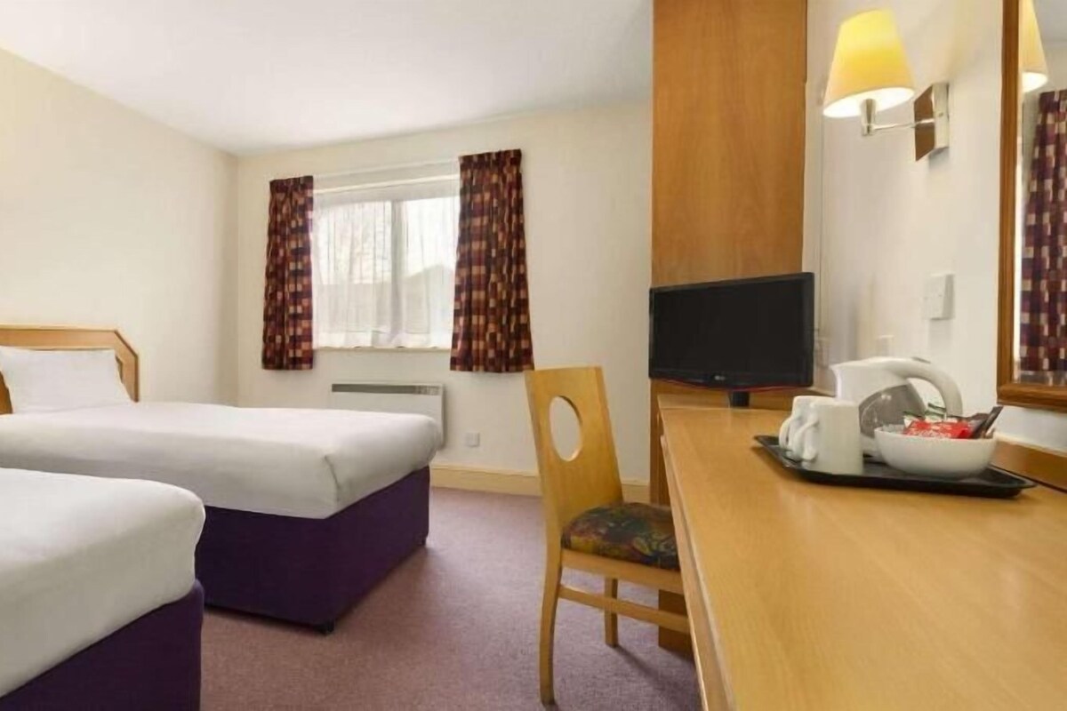 Quarto de hotel com duas camas, uma escrivaninha com televisão e uma janela com cortinas.