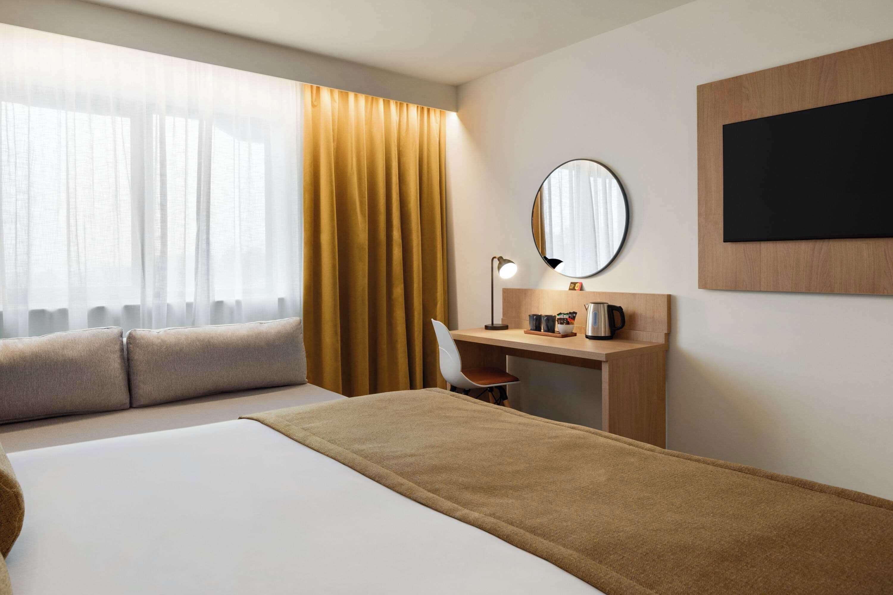 A hotel room with a bed, a desk, a chair, a desk lamp, a round mirror, and a television mounted on the wall.