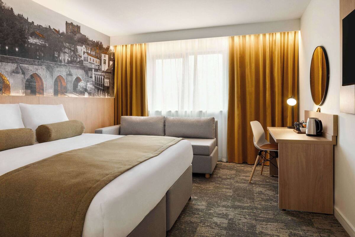 A hotel room with a bed, a desk, a chair, a large window with curtains, and a wall-mounted TV.