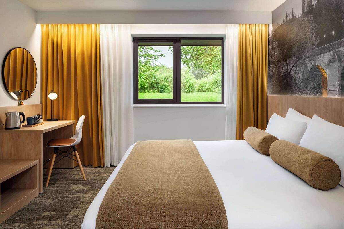 A hotel room with a bed, a desk, a chair, a window with a view of greenery, and a painting on the wall.