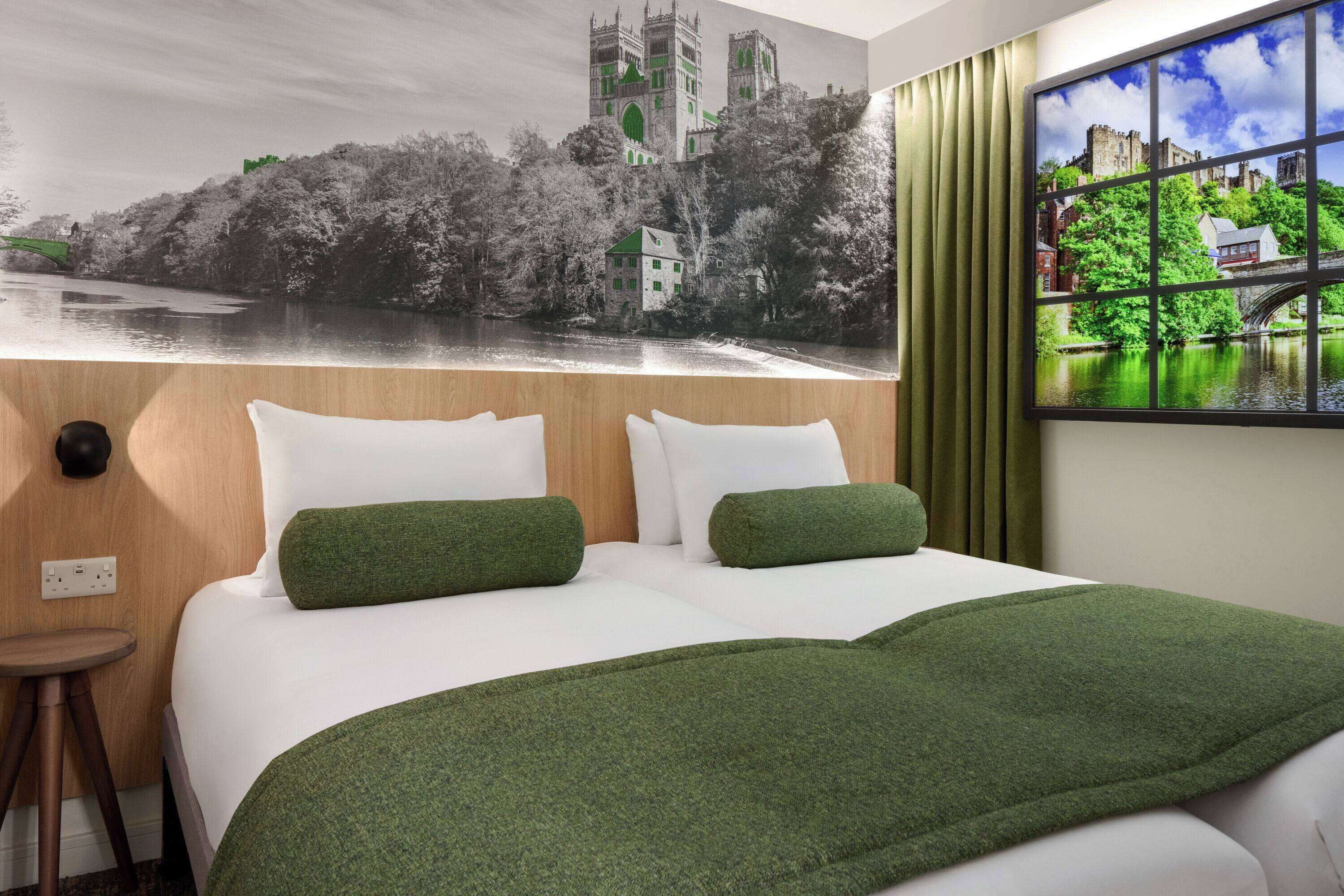 A hotel room with a large bed, two pillows, a green throw blanket, a small stool, and a wall mural depicting a castle by a river.