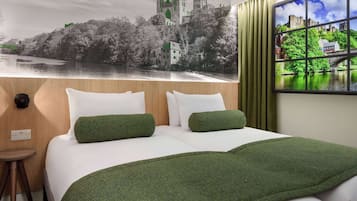 A hotel room with a large bed, two pillows, a green throw blanket, a small stool, and a wall mural depicting a castle by a river.