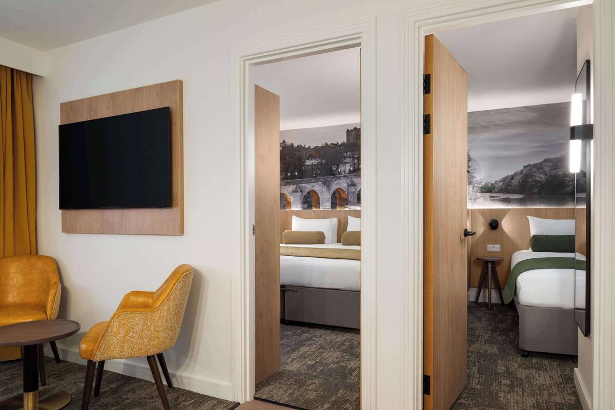 A hotel room with a bed, a chair, a TV, and a painting of a bridge.