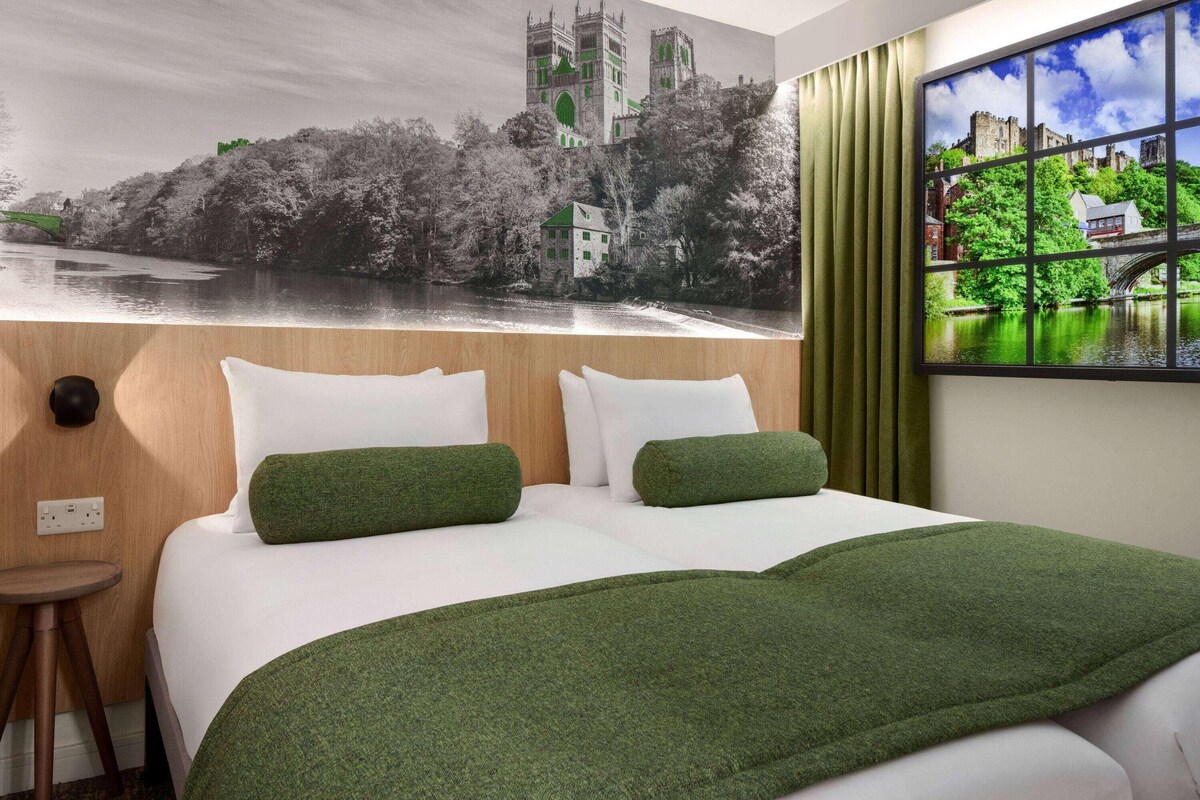 A hotel room with a bed, green pillows, a window with a view of a castle and river, and a curtain.