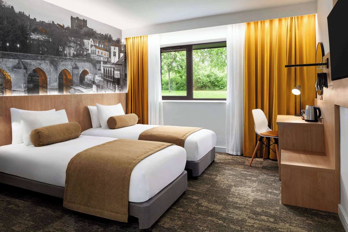 A hotel room with two beds, a desk, a chair, a window with curtains, and a mural on the wall.