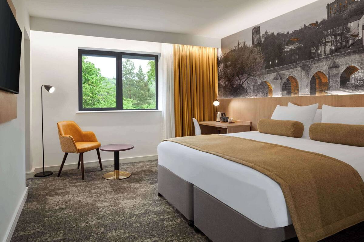 A hotel room with a large bed, a desk, a chair, a window with a view of greenery, and a cityscape mural on the wall.