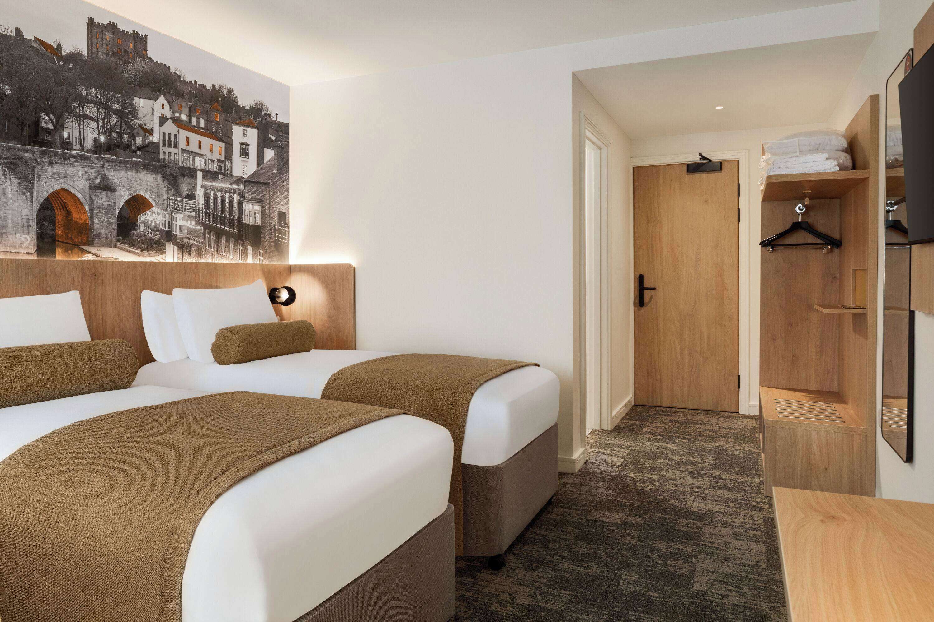 A hotel room with two beds, a wooden wardrobe, a mirror, and a painting of a town on the wall.