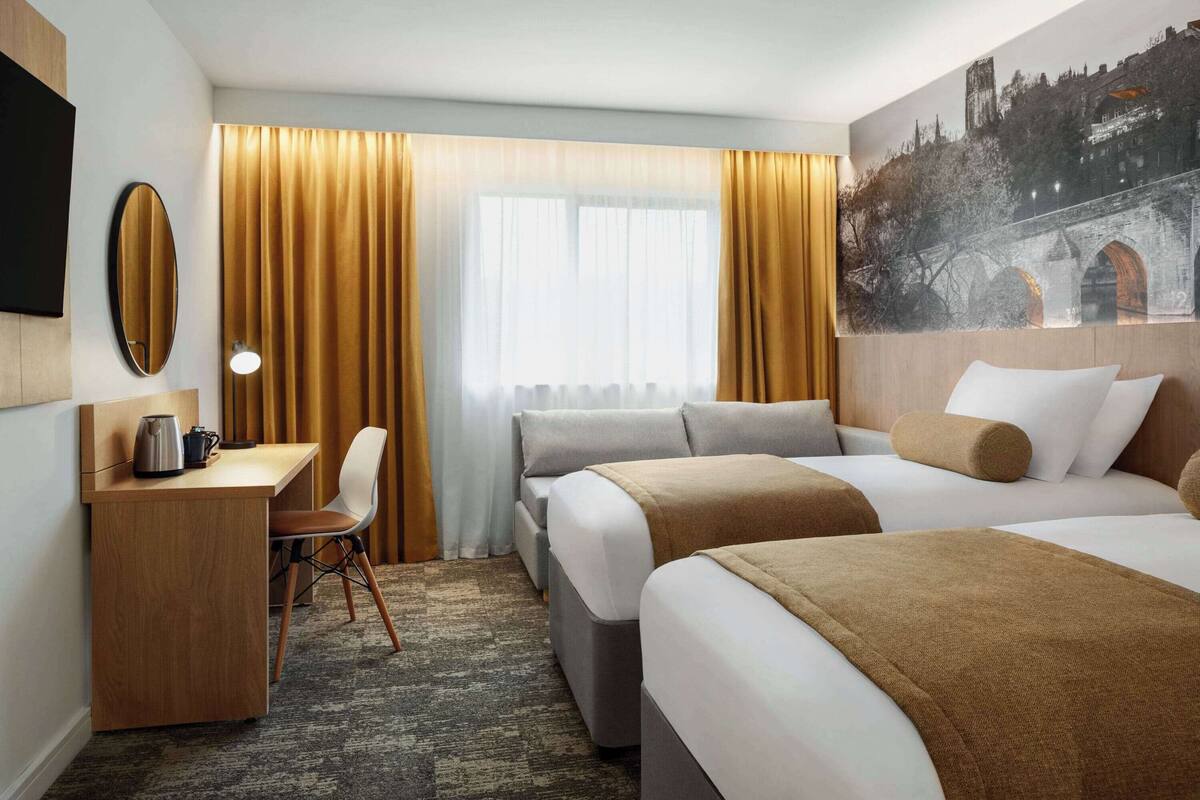 A hotel room with a desk, two beds, a chair, and a large window with curtains.