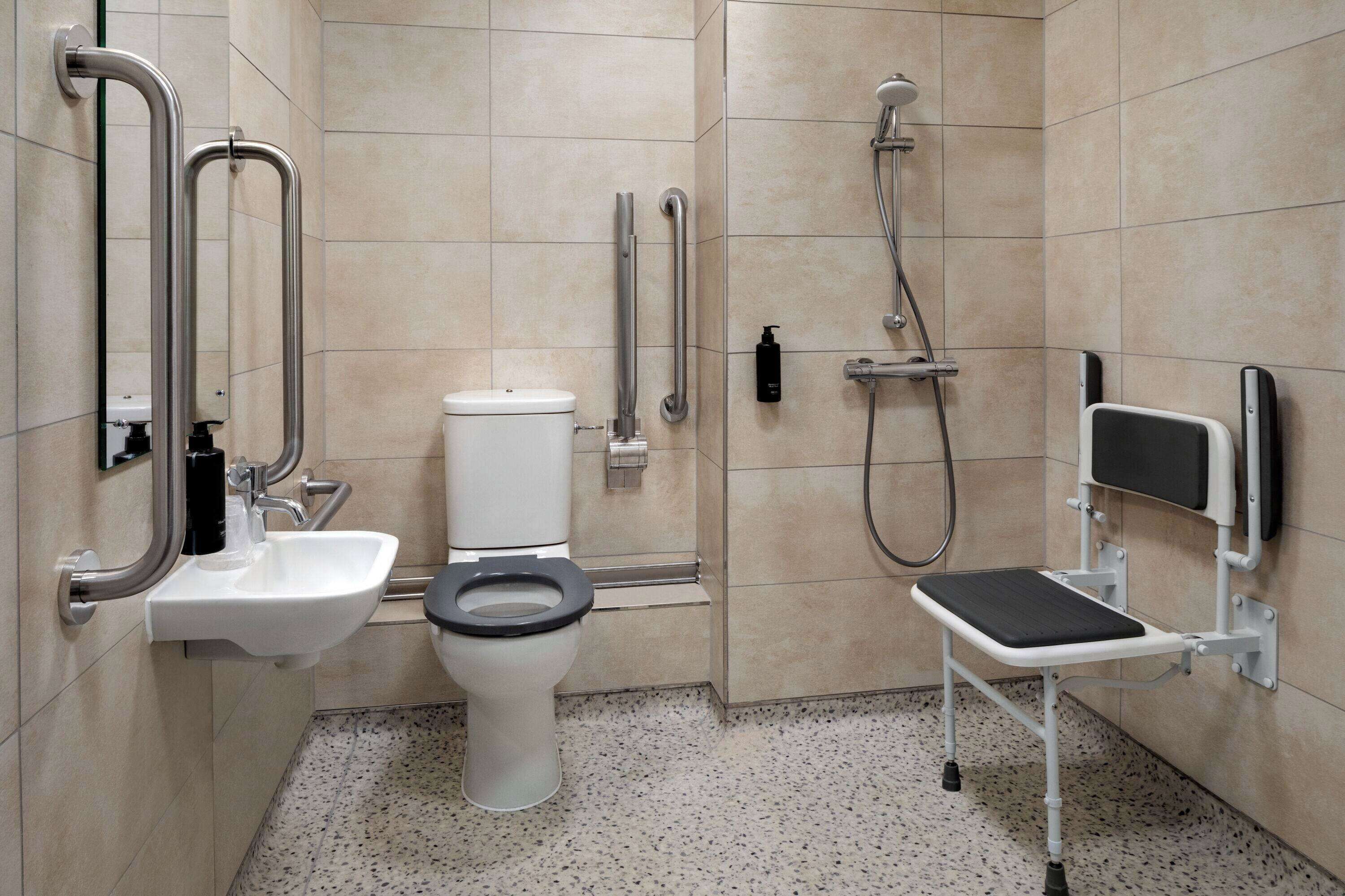 A bathroom with a toilet, sink, and shower. It includes grab bars and a wheelchair-accessible bench.