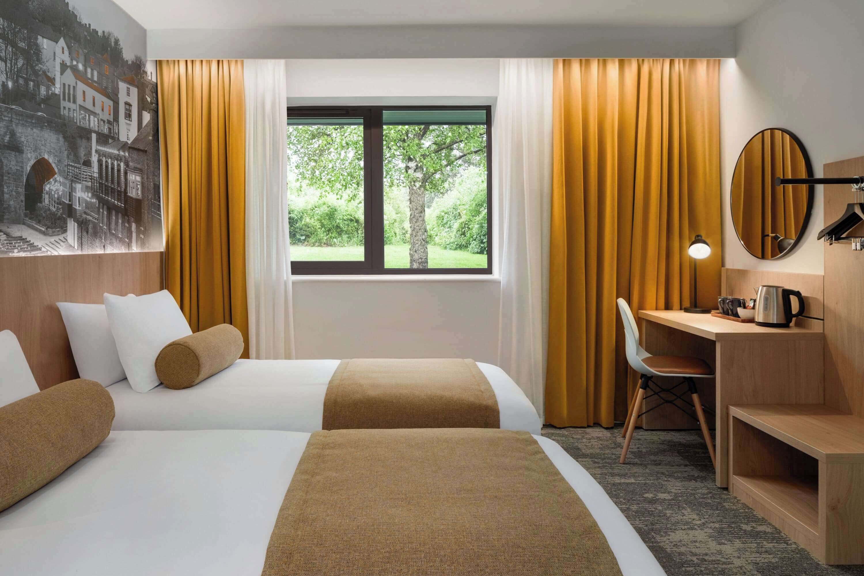 Une chambre d’hôtel avec deux lits, un bureau, une chaise, une grande fenêtre avec des rideaux et un miroir fixé au mur.