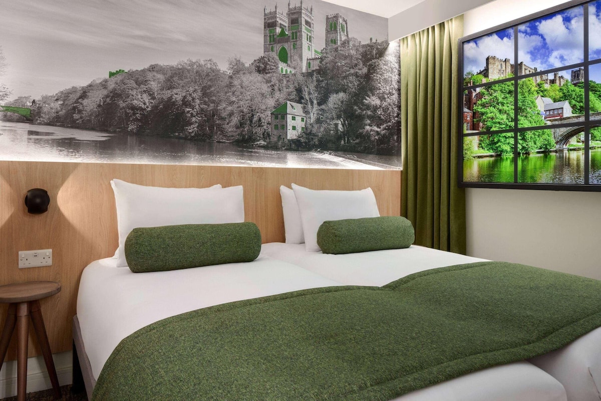 Une chambre d’hôtel avec un lit, des oreillers verts, une fenêtre donnant sur un château et une rivière, et un rideau.