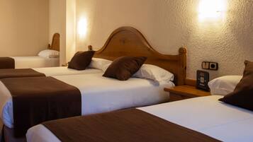 Habitación de hotel con tres camas individuales, cada una con cabecero de madera, ropa de cama blanca y almohadones marrones. Se observa una mesita de noche y un reloj en la pared.