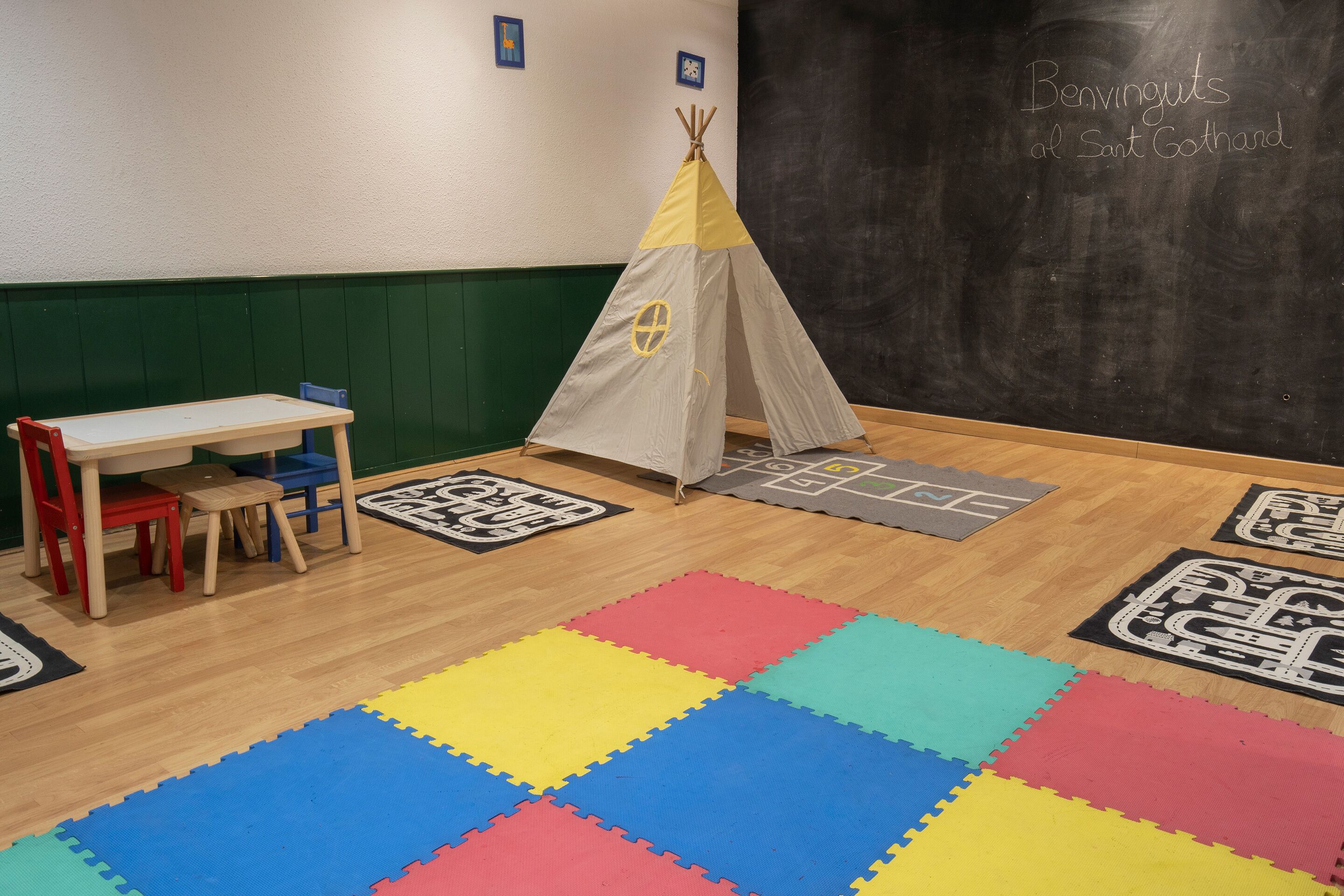Una habitación con piso de madera, alfombras de espuma coloridas, una mesita con sillas, un tipi y una pizarra con escritura.