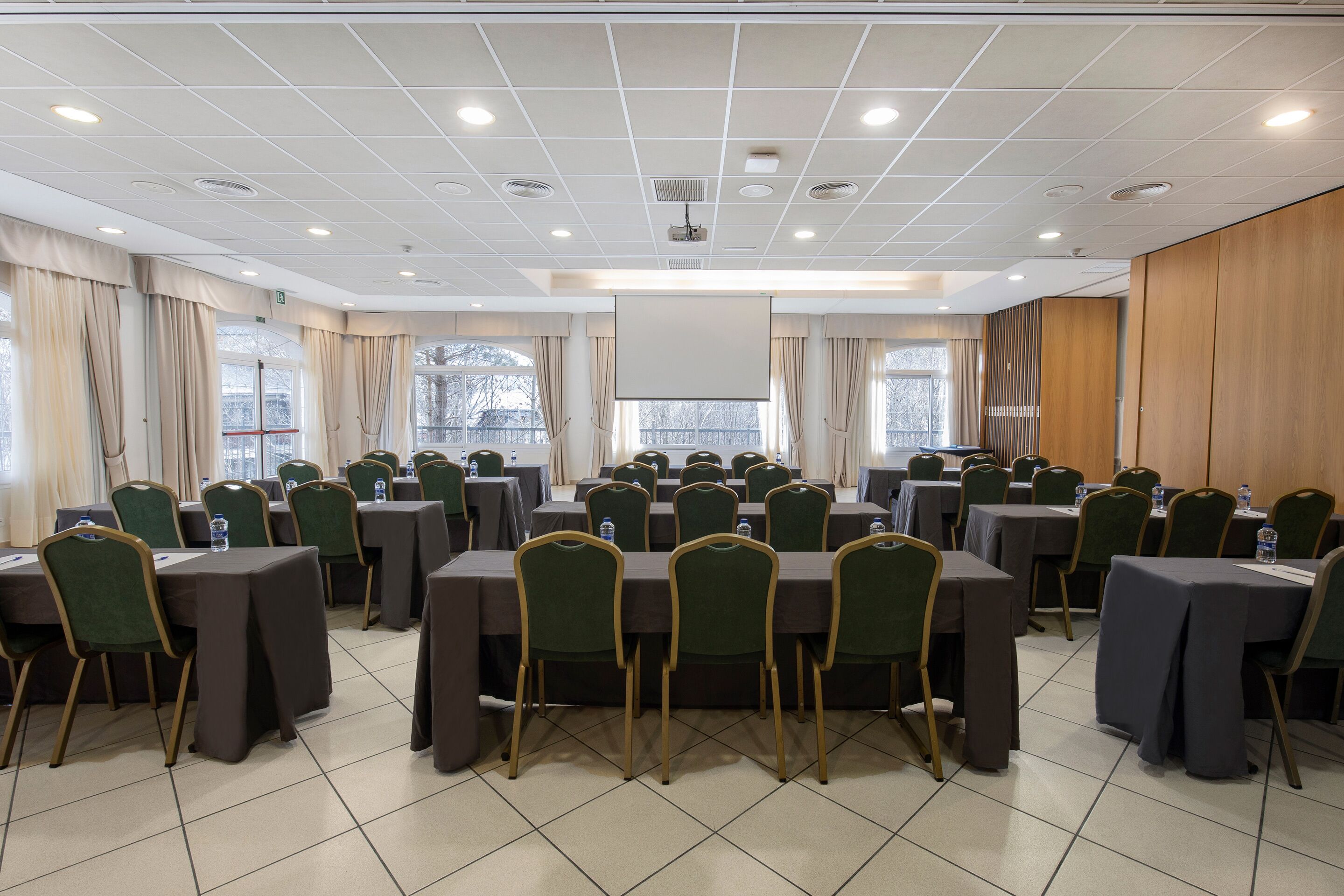 Un salón de conferencias con varias mesas y sillas, una pantalla de proyector y ventanas grandes con cortinas.