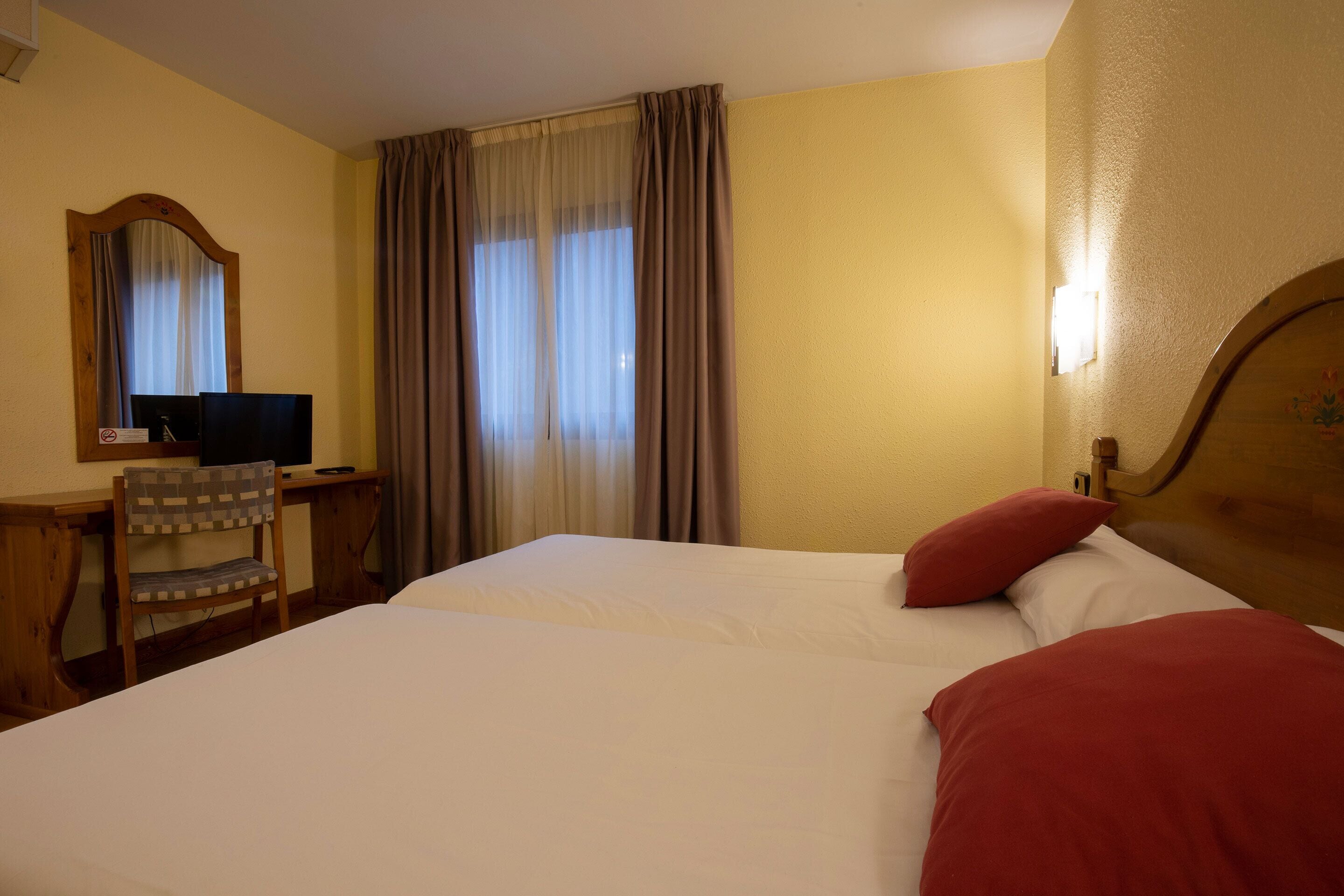 A hotel room with a bed, a desk with a computer, a chair, and a window with curtains.