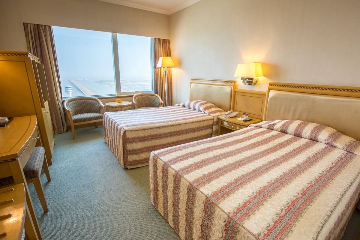 A hotel room with two beds, a desk, a chair, and a view of the city.