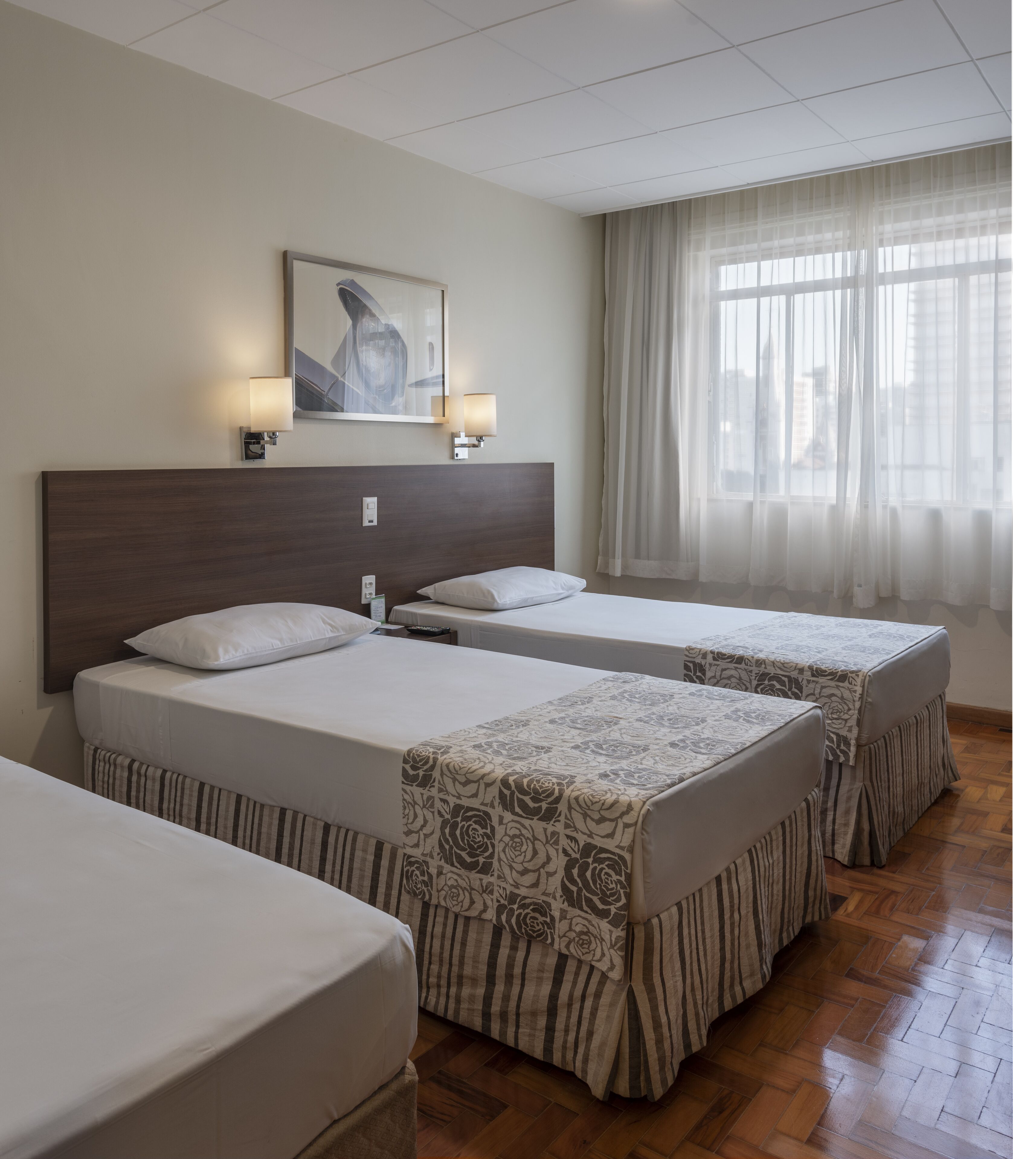 A hotel room with two beds, a wooden headboard, a painting on the wall, and a window with sheer curtains.