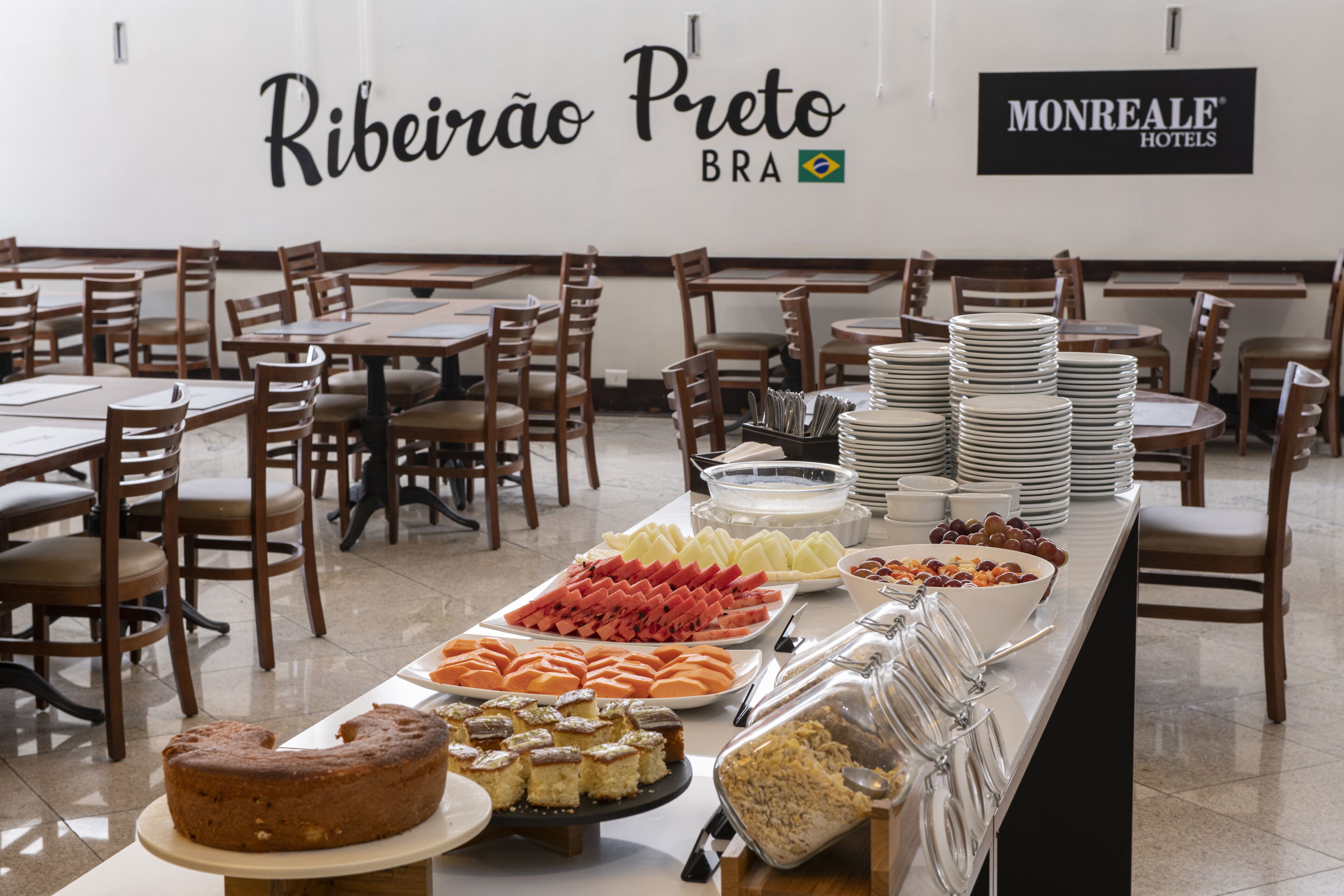 Buffet montado com diversos alimentos e um bolo sobre uma mesa, rodeado por cadeiras e mesas em uma área de refeições.