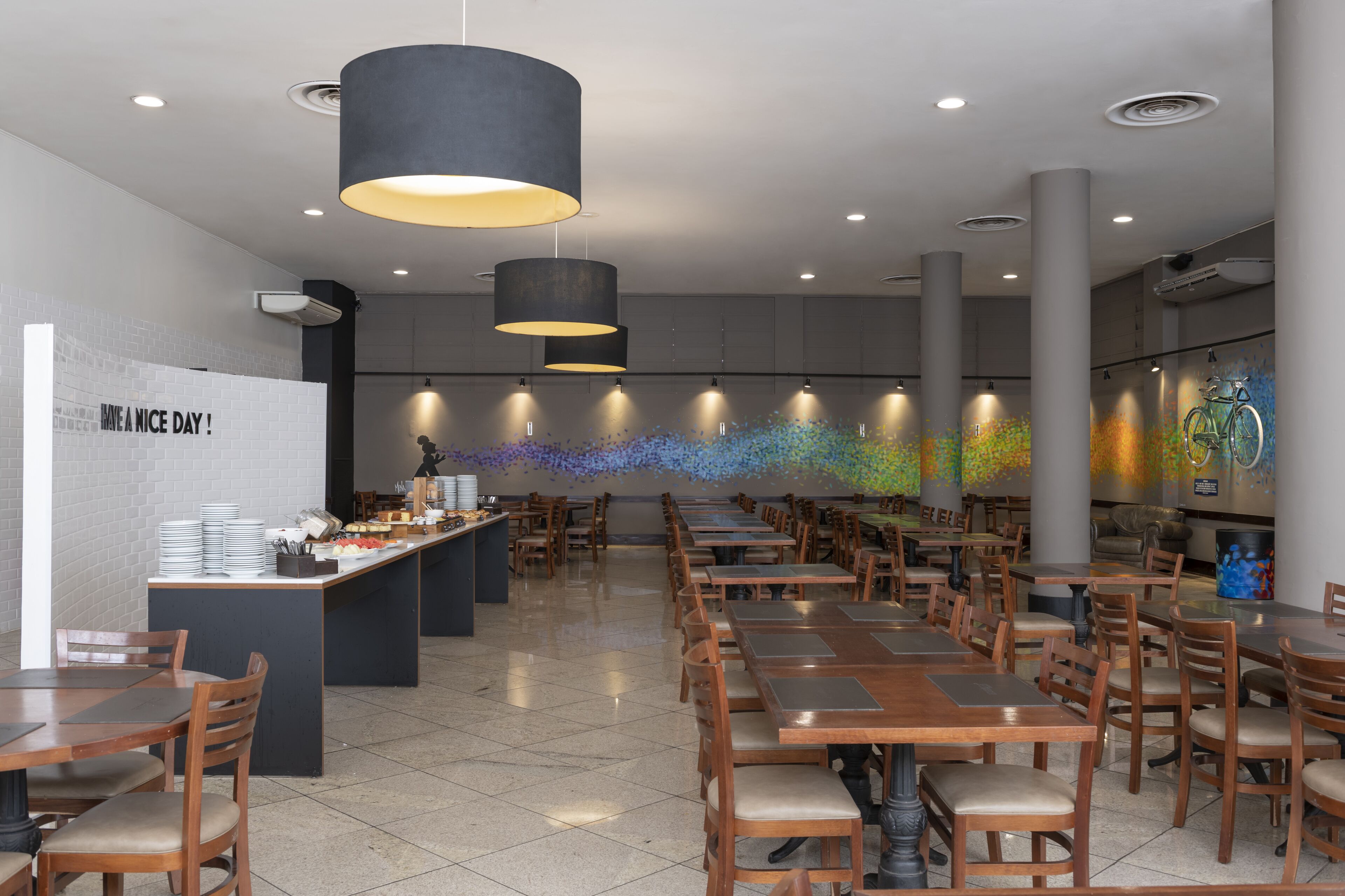 Un'ampia sala da ristorante con numerosi tavoli e sedie, un'isola centrale con servizio buffet e un colorato murale sulla parete di fondo.