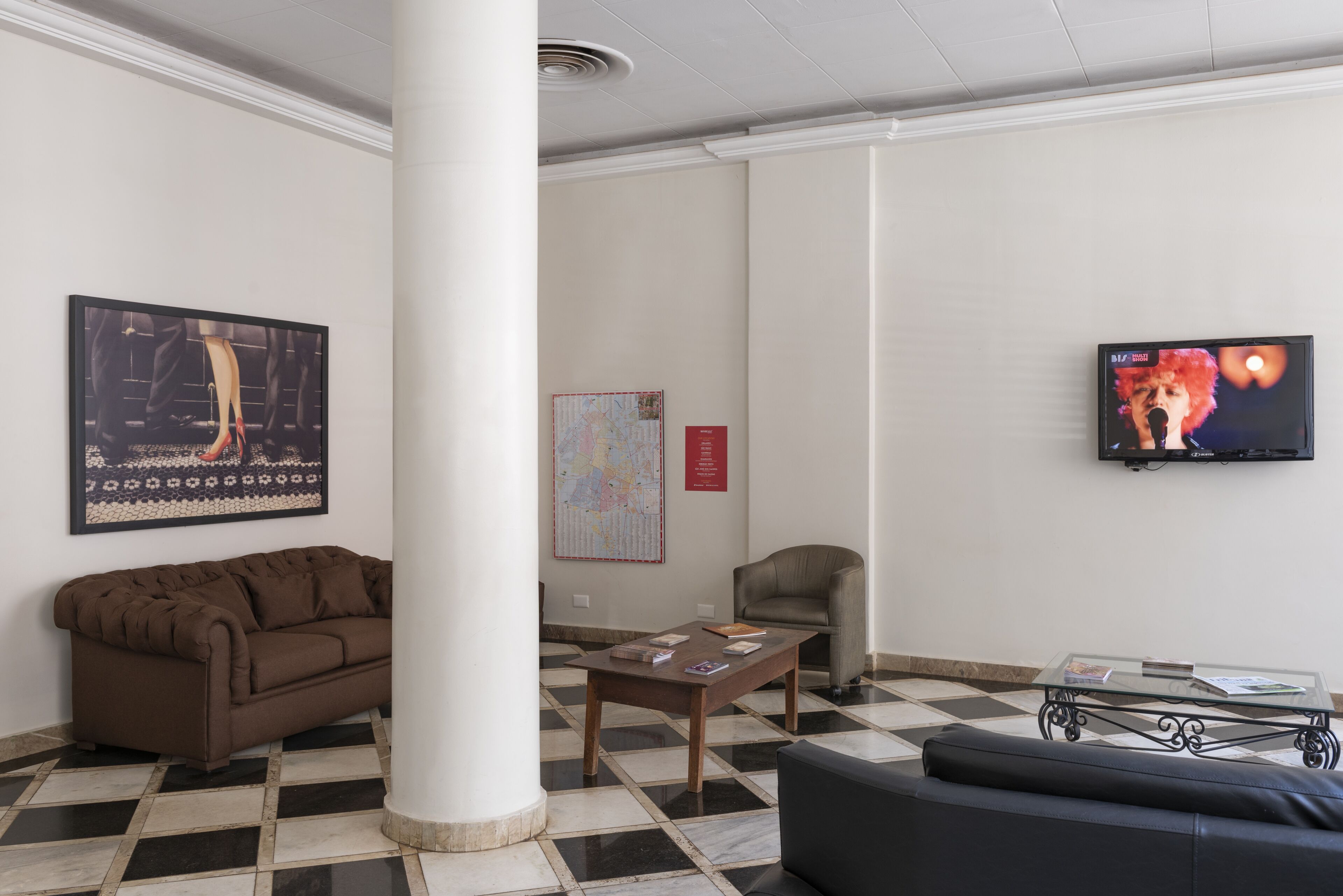 A lobby with a checkered floor, a brown sofa, a glass coffee table, a map on the wall, and a television displaying a person.