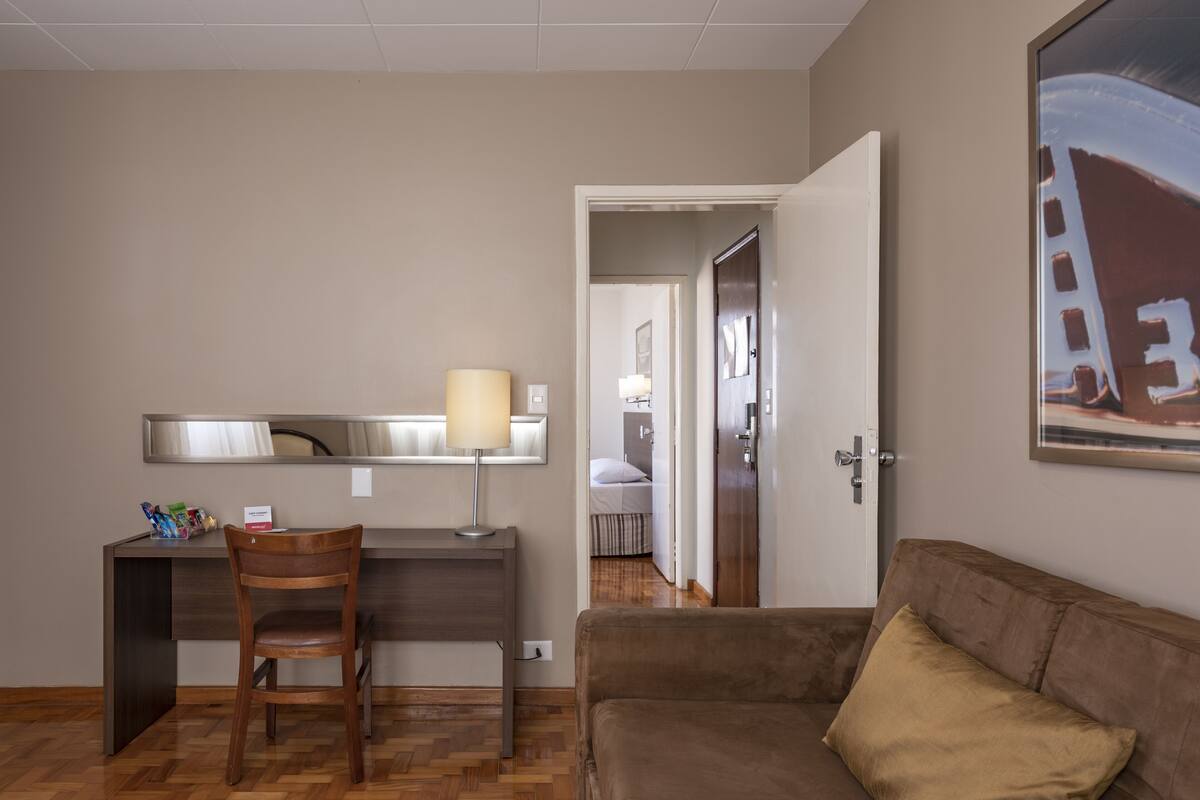 Ein Hotelzimmer mit einem Holztisch, einem Sessel, einer Lampe, einem braunen Sofa und einem Bild an der Wand.