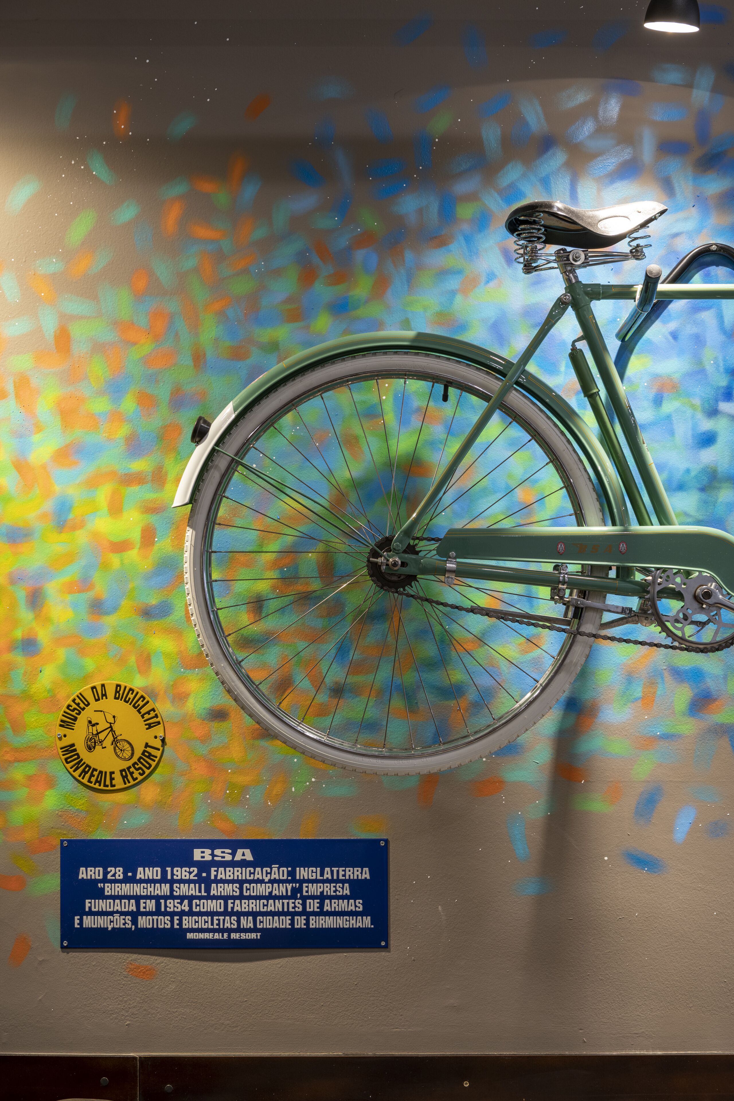 A vintage bicycle with a chain and seat, mounted on a wall with a colorful abstract background. A plaque with text and a logo is visible below the bicycle.