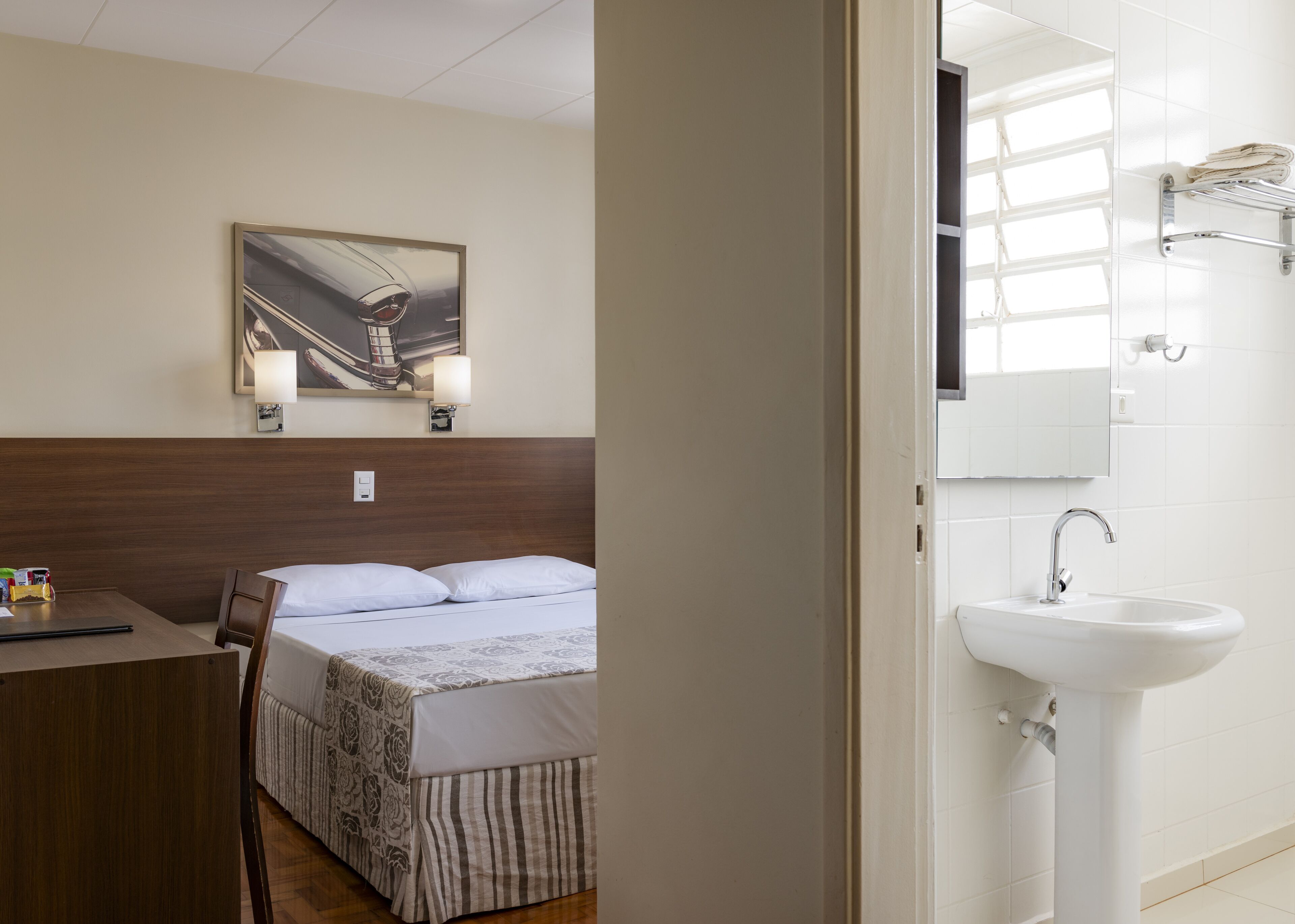 A hotel room with a bed, a desk, a chair, a painting on the wall, a sink, and a mirror.