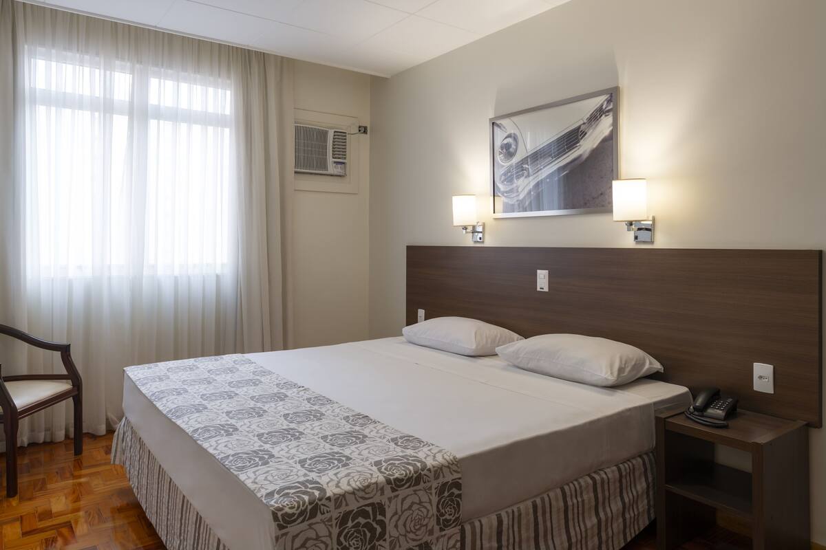 Ein Hotelzimmer mit einem großen Bett, zwei Nachttischlampen, einem Telefon, einem Sessel und einem Fenster mit Vorhängen.