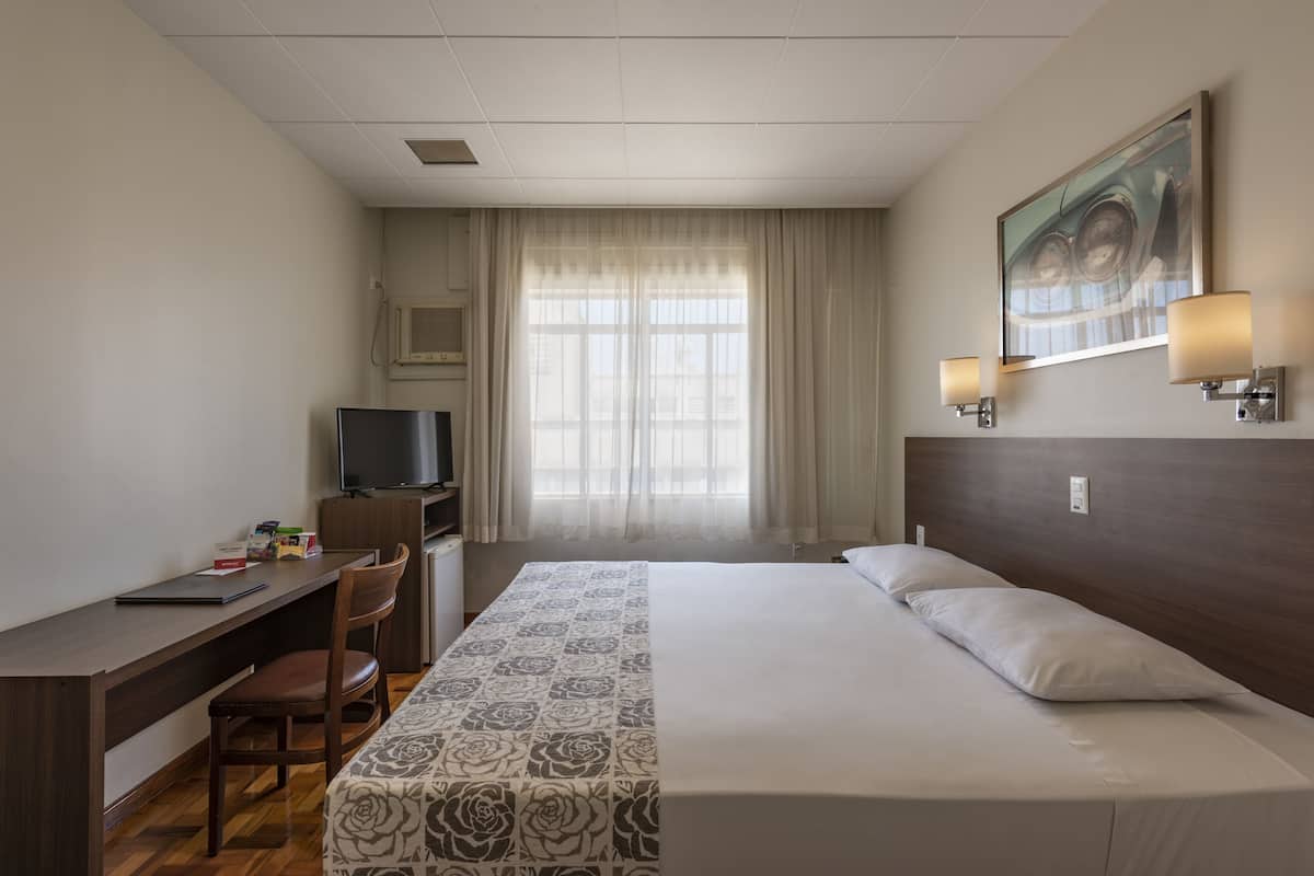 Ein Hotelzimmer mit einem Bett, einem Schreibtisch, einem Stuhl, einem Fernseher und einem Fenster mit Vorhängen.