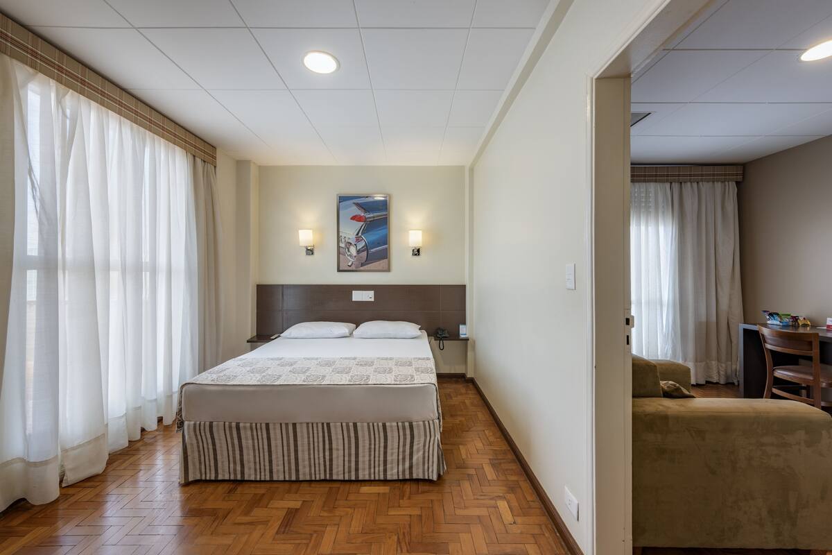 Ein Hotelzimmer mit einem großen Bett, einem Schreibtisch und einem Sessel. Zwei Wandlampen und ein Gemälde sind an der Wand angebracht.