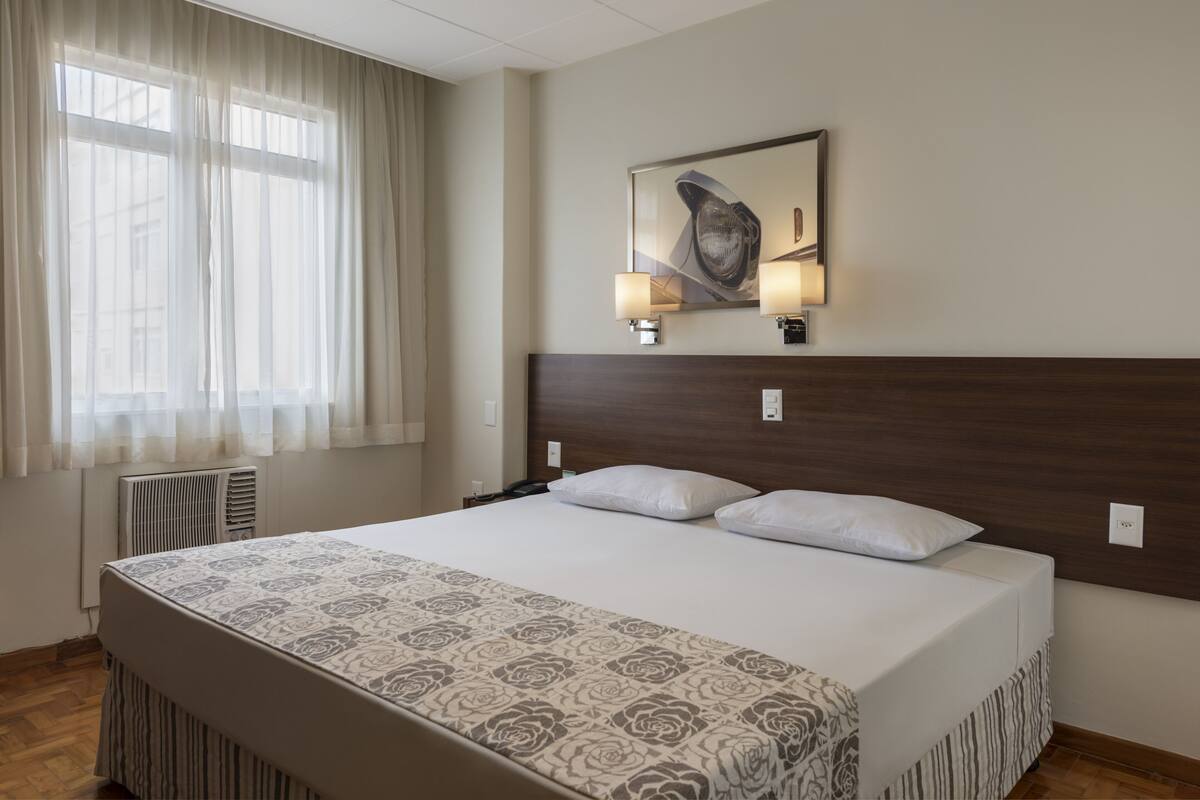Ein Hotelzimmer mit einem großen Bett, einem Kopfteil mit Wandleuchten, einem Heizkörper und einem Fenster mit durchsichtigen Vorhängen.