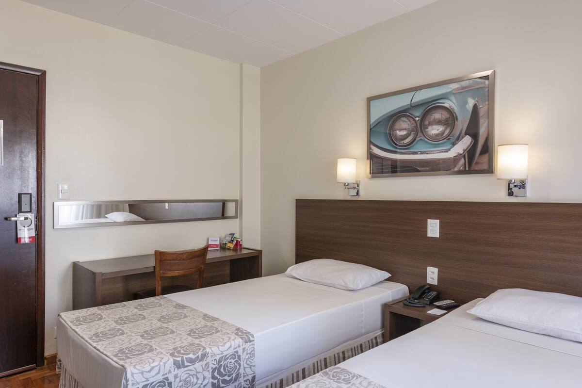 Ein Hotelzimmer mit zwei Einzelbetten, einem Schreibtisch, einem Stuhl, einem kleinen Tisch, einem Telefon und einem Bild an der Wand.