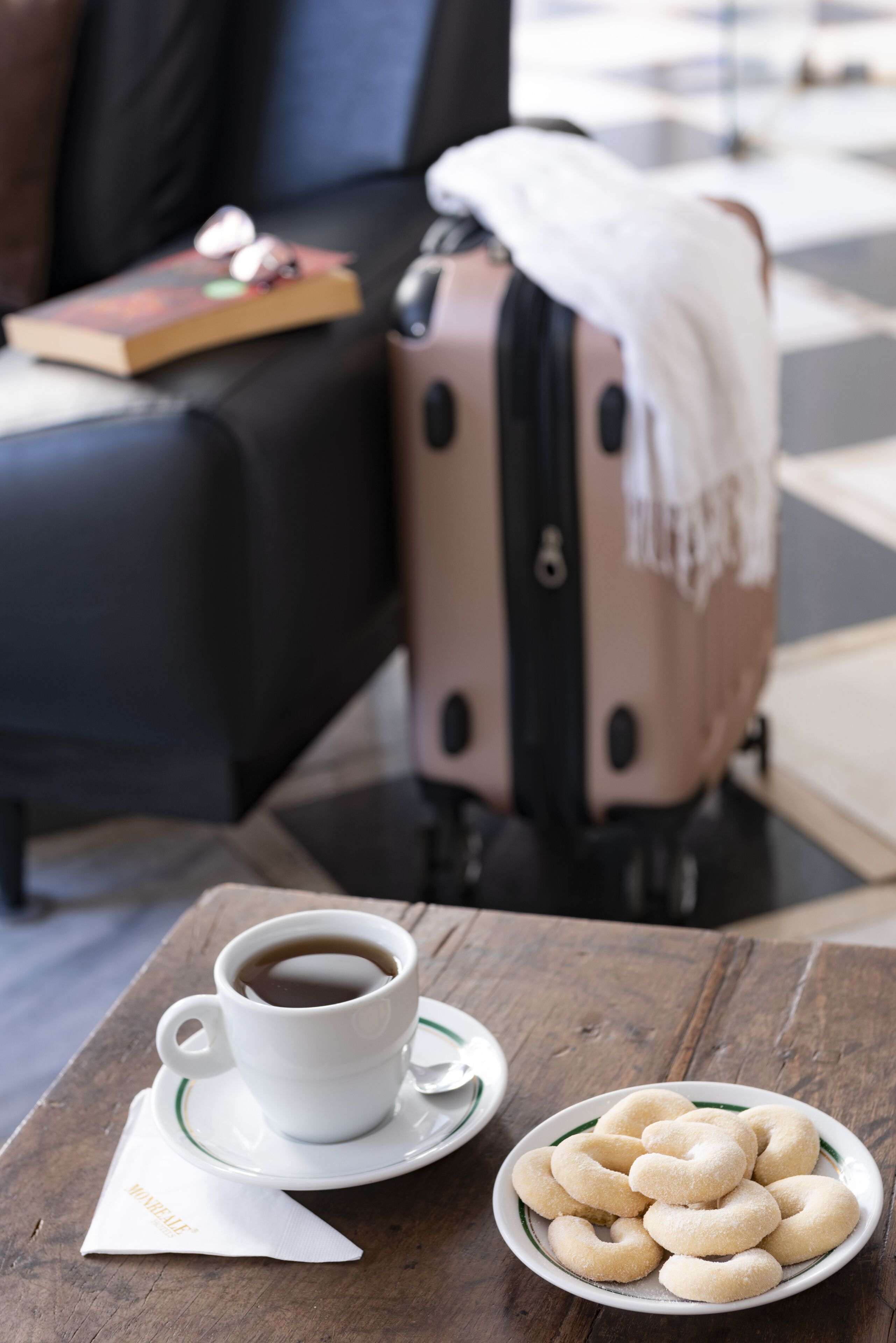 A table with a cup of coffee, a plate of cookies, a suitcase, and a book on a couch.