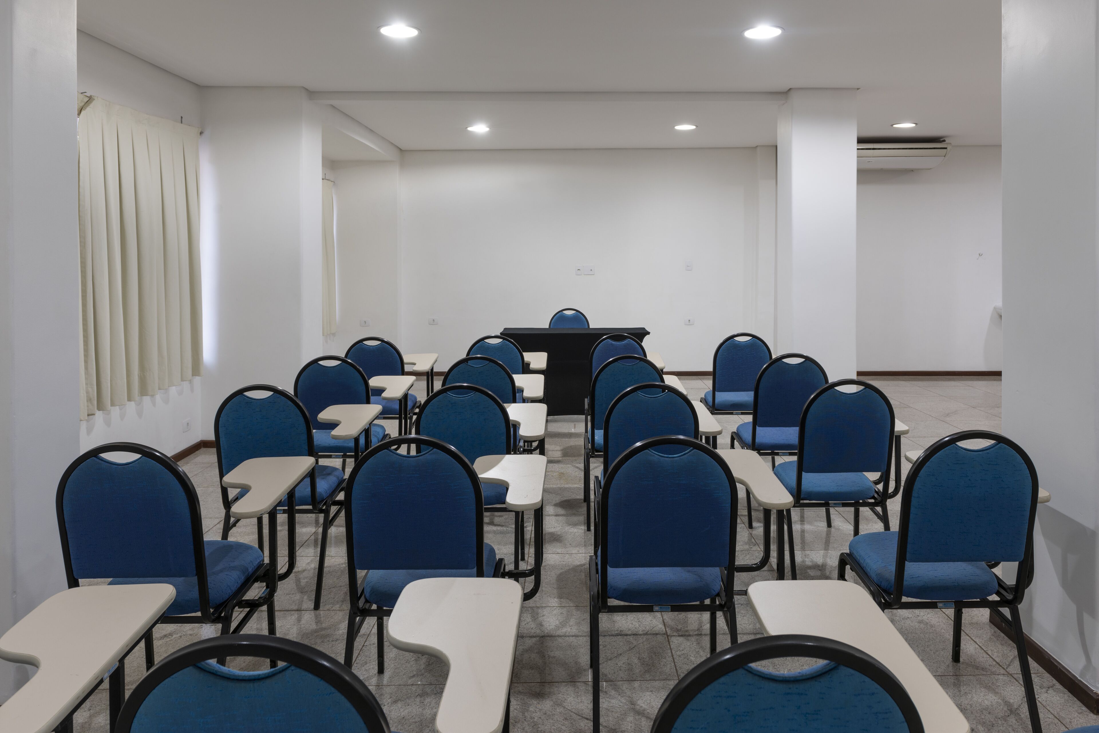 A conference room with rows of blue chairs and white tables, a podium at the front, and a window with blinds.