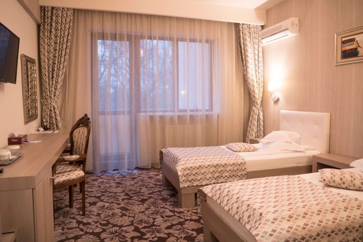 Een hotelkamer met twee bedden, een bureau, een stoel en een groot raam met dunne gordijnen.