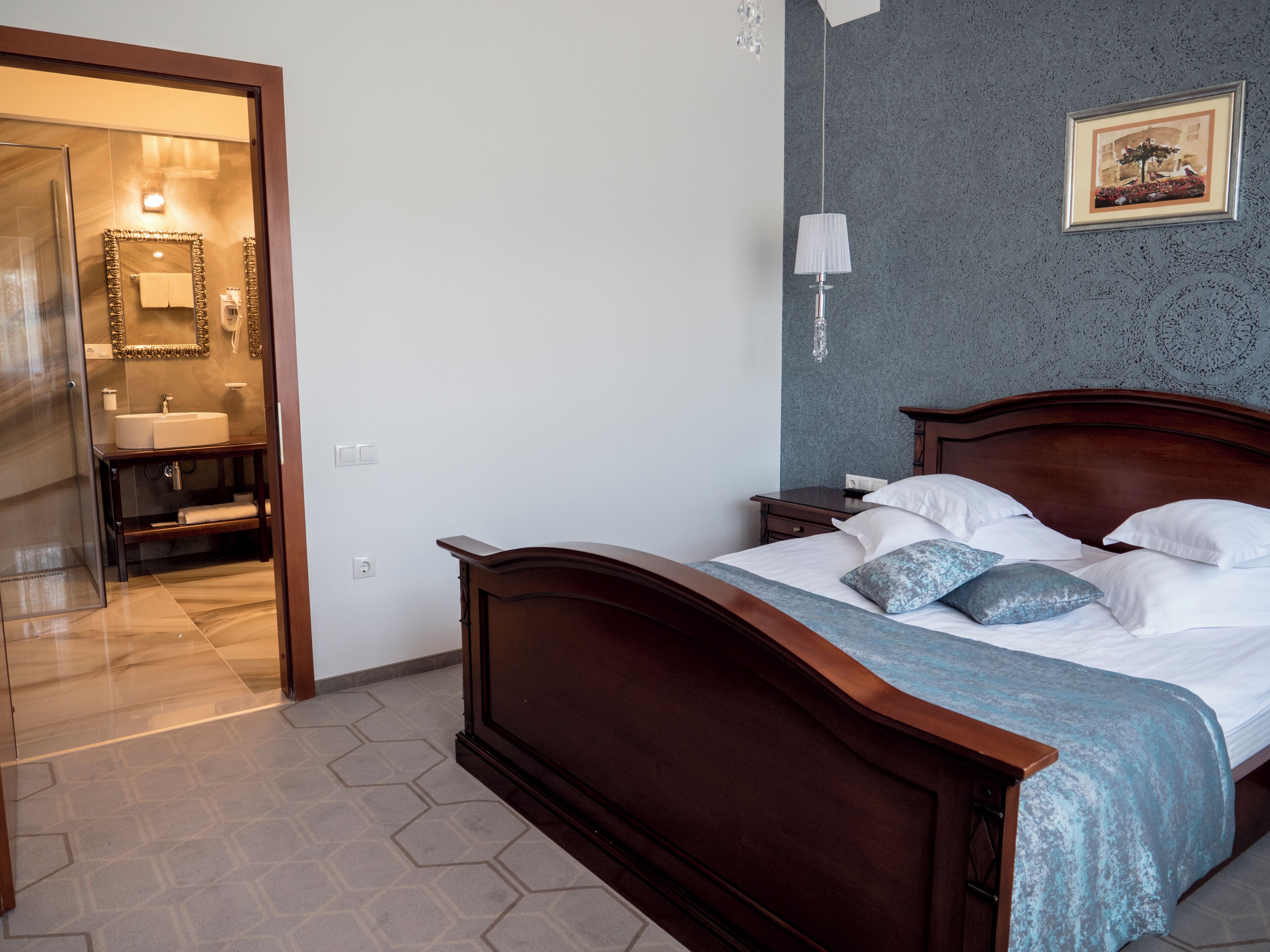 A bedroom with a wooden bed, a nightstand, a wall-mounted lamp, and a bathroom visible through an open door.