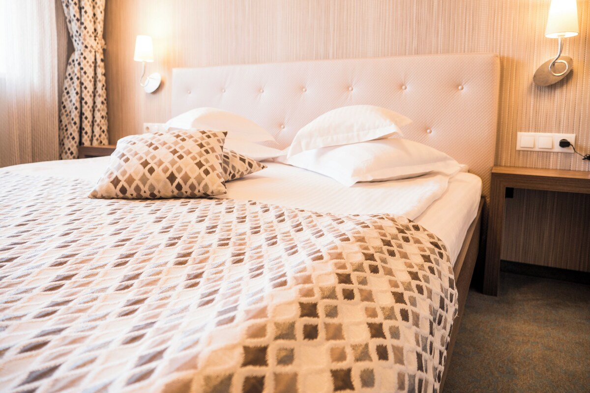 Een net opgemaakt bed met een geborduurd sprei, twee kussens en een hoofdbord. Er zijn twee nachtkastjes met lampen en een stopcontact op de muur.