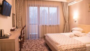 Habitación de hotel con una cama, un escritorio y una silla. Se ve una ventana con cortinas, un equipo de aire acondicionado y un televisor montado en la pared.