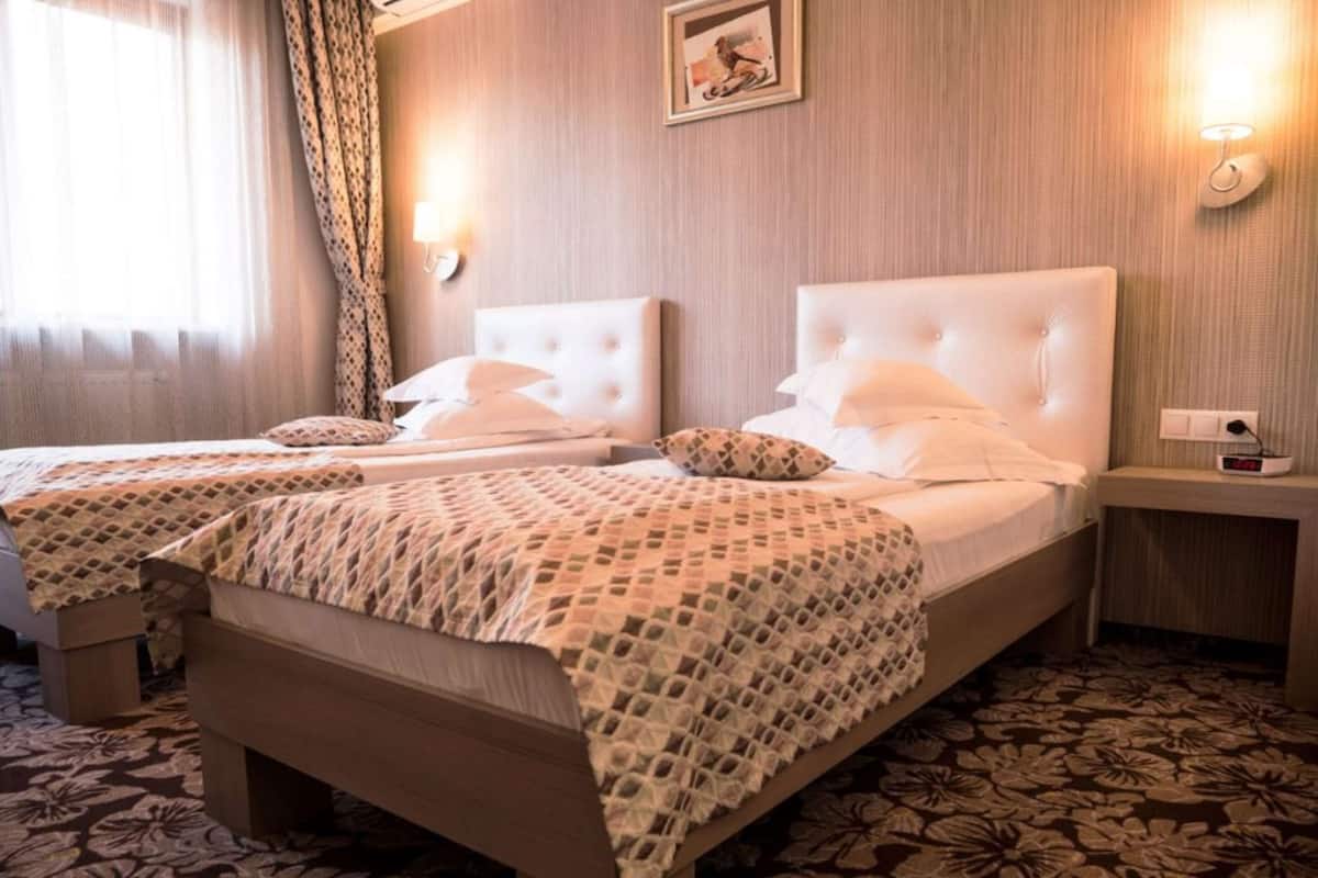 A hotel room with two beds, a bedside table, a clock, and a wall-mounted light.